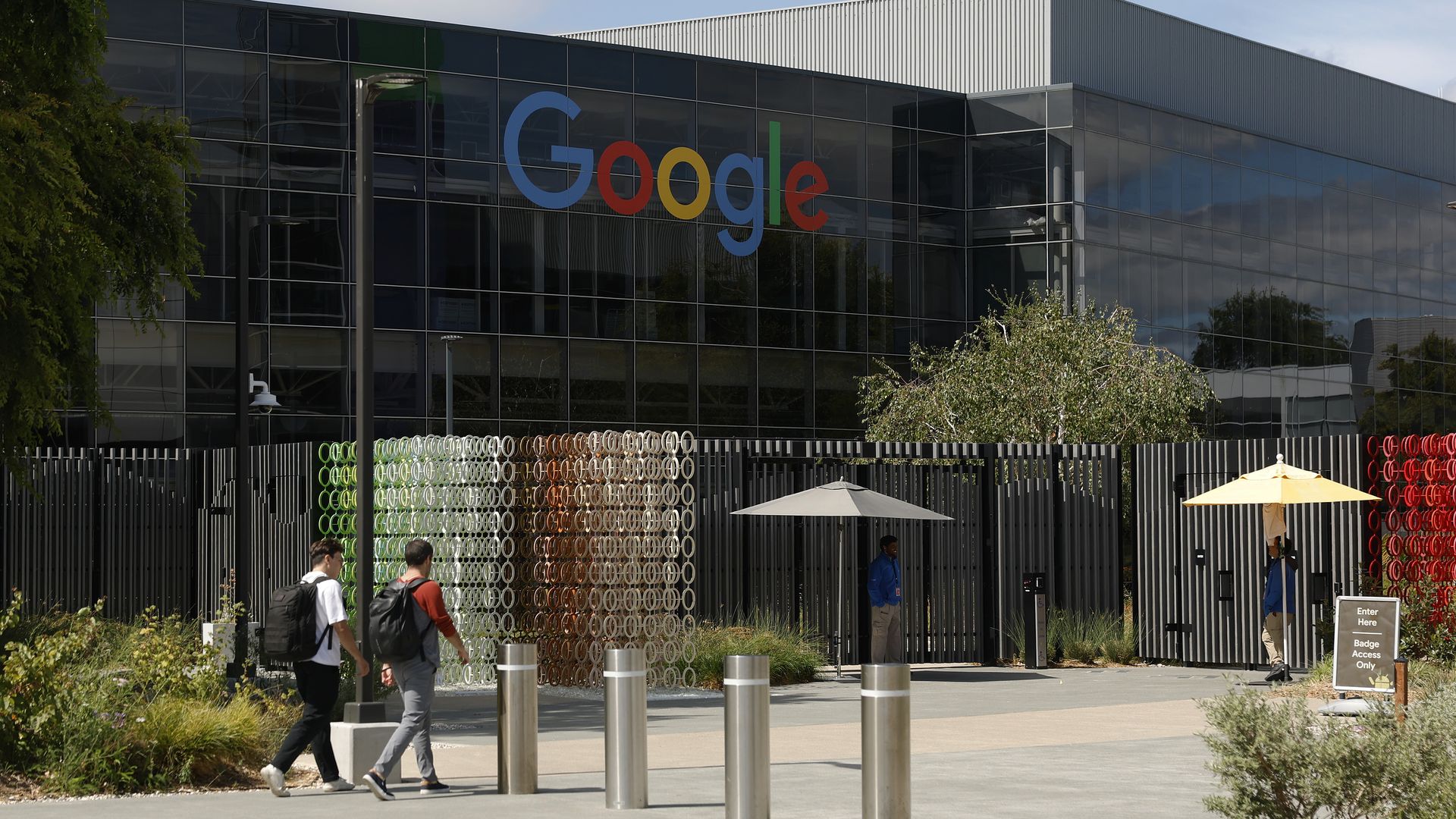 Google workers, one wearing a white tee-shirt and black pants and the other a red sweater and gray jeans, both with black backpacks, enter a building on the Google headquarters campus, which features the Google logo at the top. Vegetation can be seen outside.