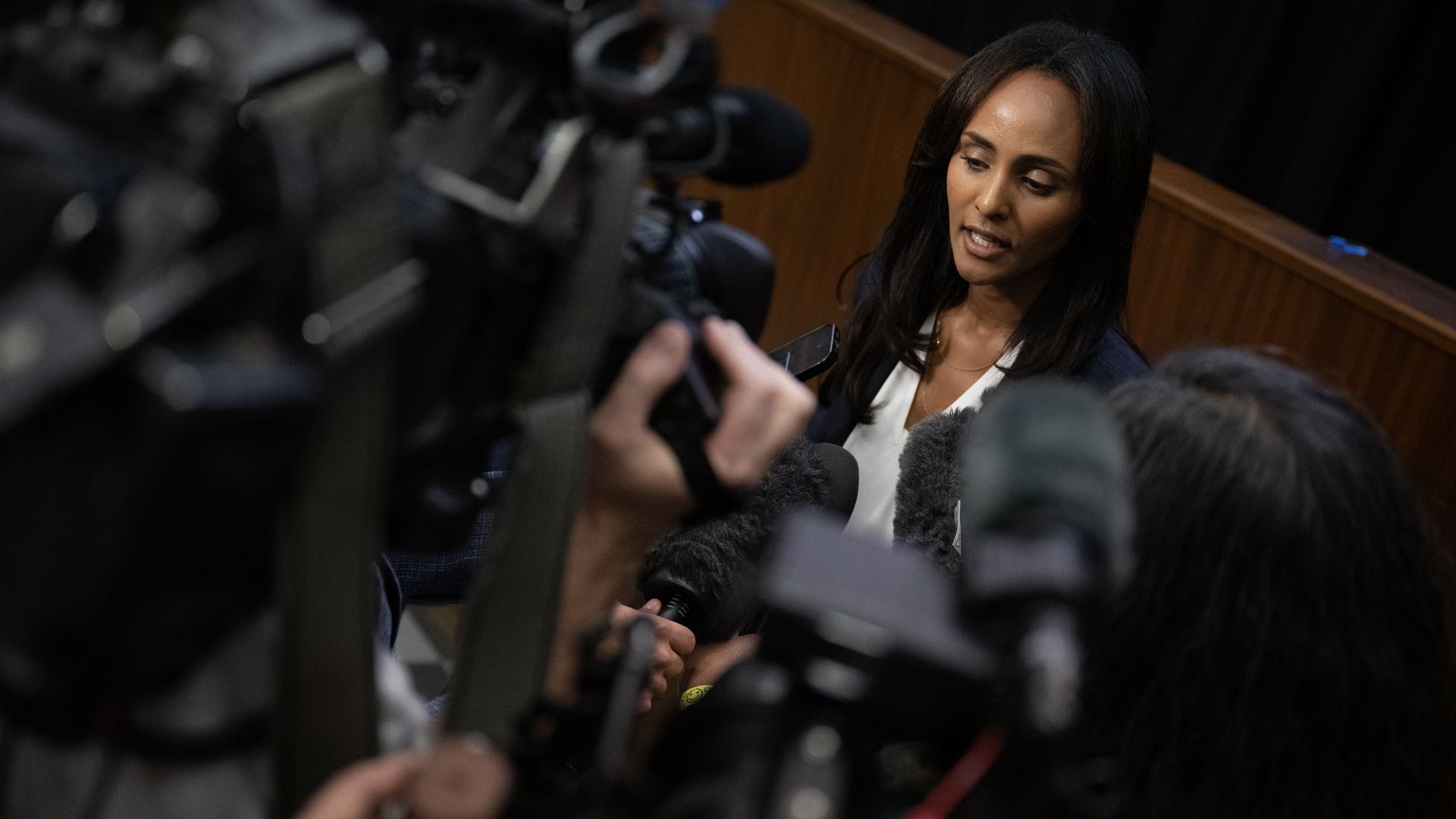 A woman with long dark hair speaks to reporters at a press conference, surrounded by microphones and cameras; blurred foreground gear and a wooden-panel backdrop.