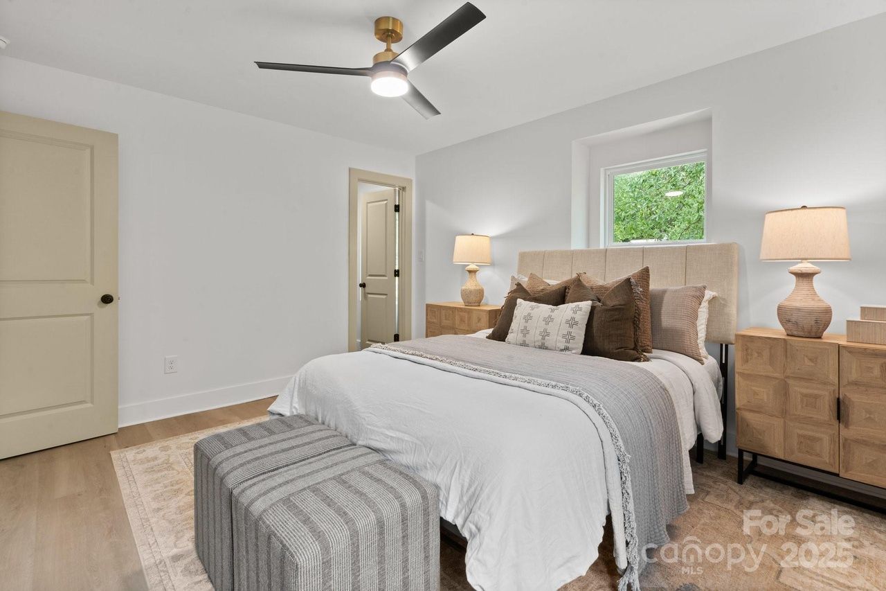 Minimalist bedroom with white walls, beige door and headboard, wooden nightstands with lamps, bed with white and gray bedding and assorted brown and white pillows, striped gray bench, and ceiling fan.