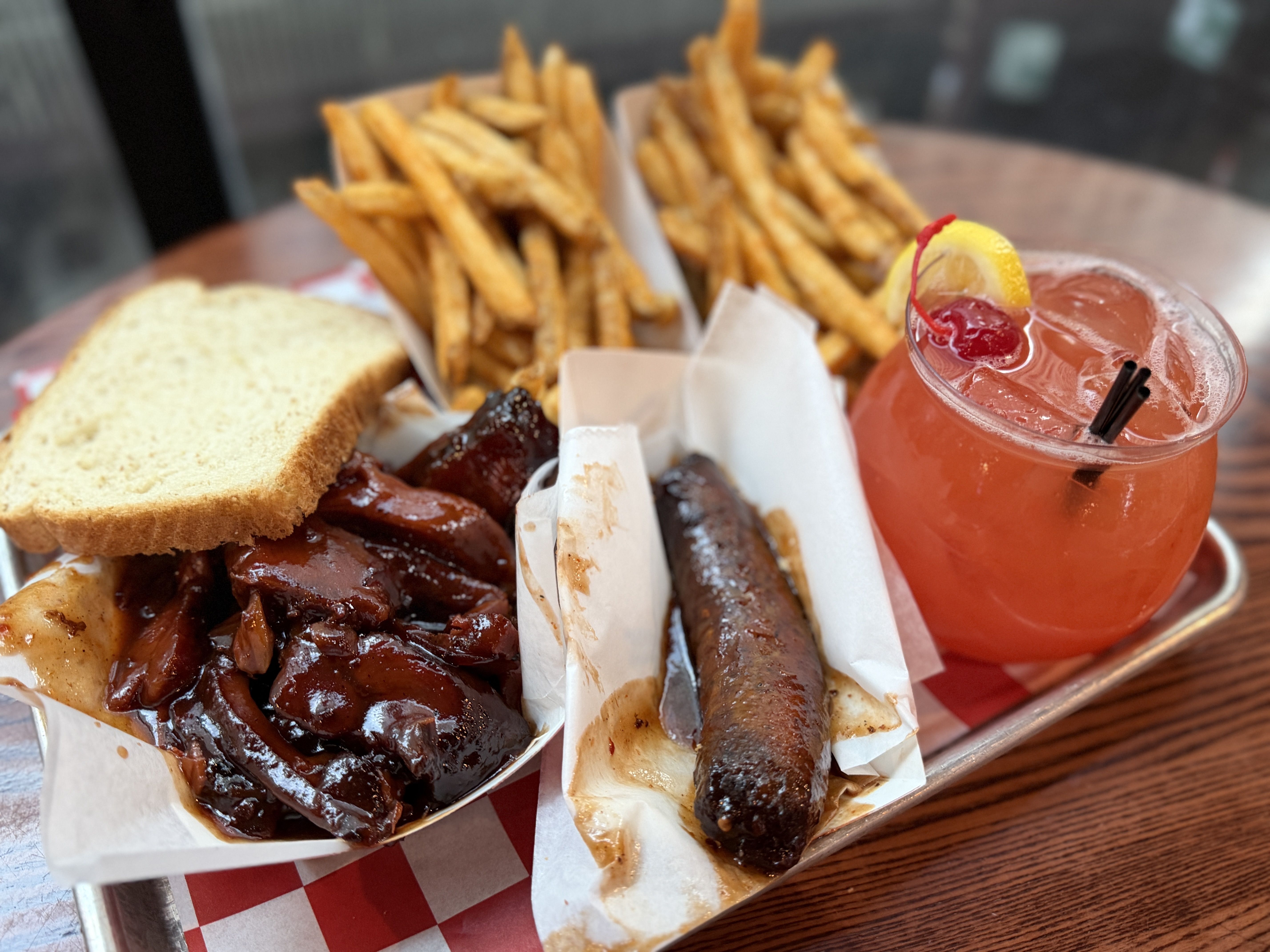 Metal tray with glazed barbecue ribs, a grilled sausage, toast, and crispy fries. A red cocktail with lemon wedge and cherry sits beside on checkered paper.
