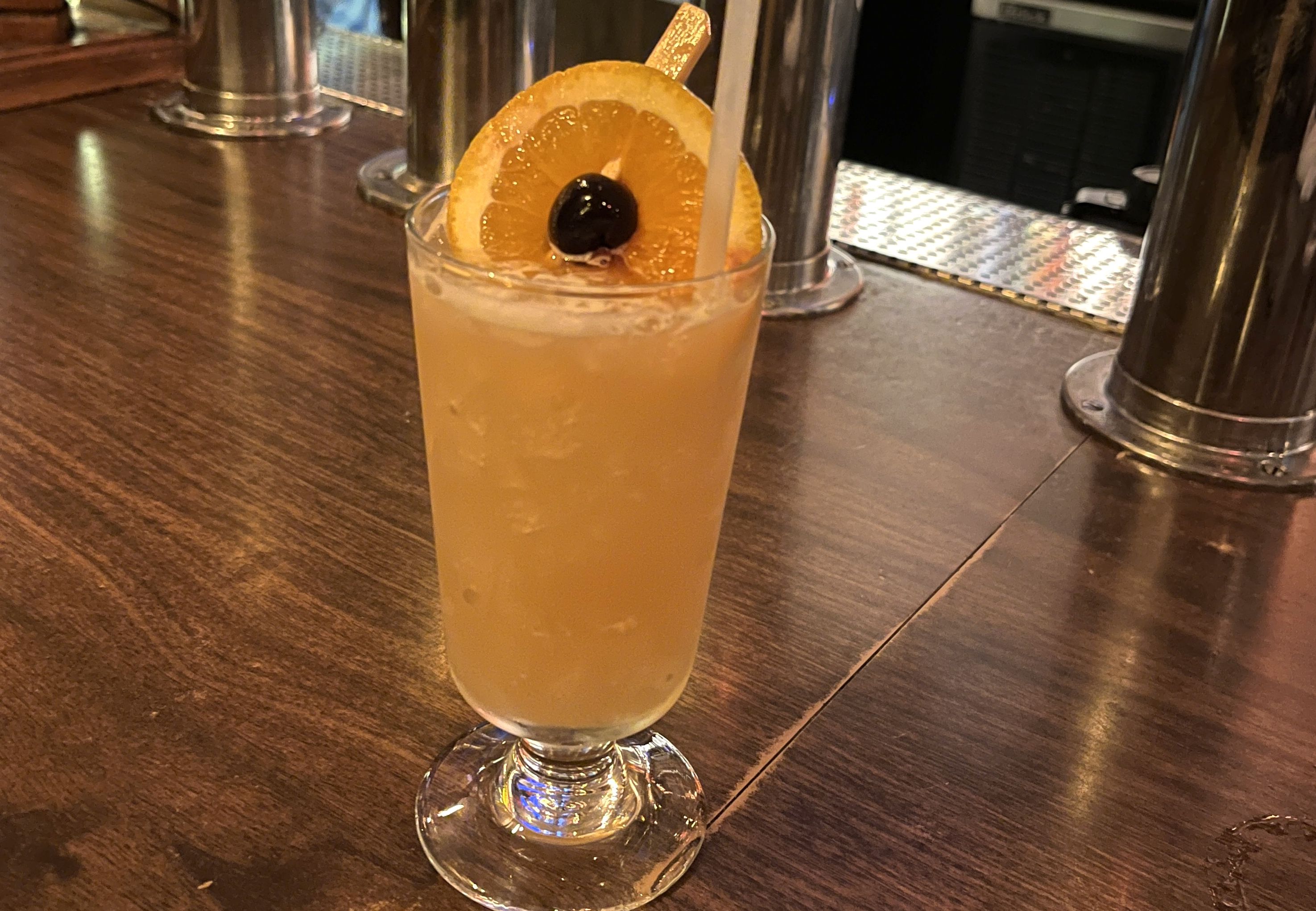 Orange-yellow cocktail in a tall glass with crushed ice, garnished with an orange slice and a dark cherry, on a wooden bar counter with metal fixtures in the background.
