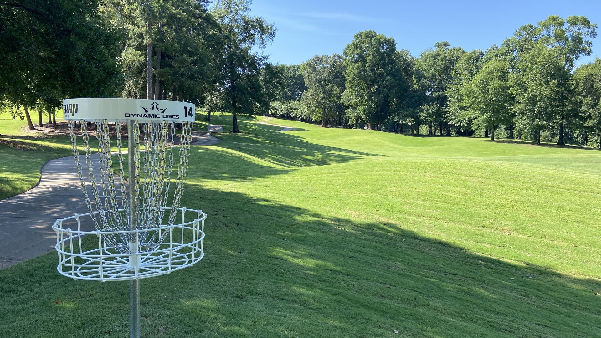 Disc golf basket with metal chains and a white rim labeled "Dynamic Discs 14" stands on a sunlit green park fairway, with a winding path, trees, and a bright blue sky in the background.