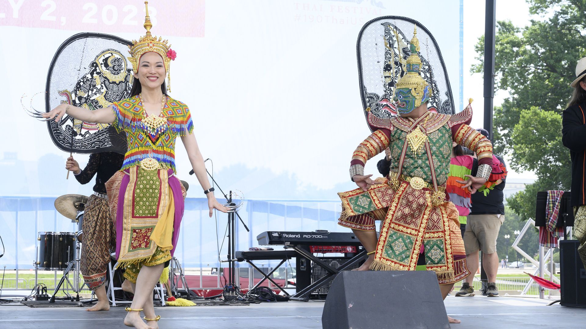 Two performers in vibrant traditional Thai costumes dancing on stage at an outdoor festival, with one wearing a green and red outfit and the other in colorful patterned attire and a gold headdress.