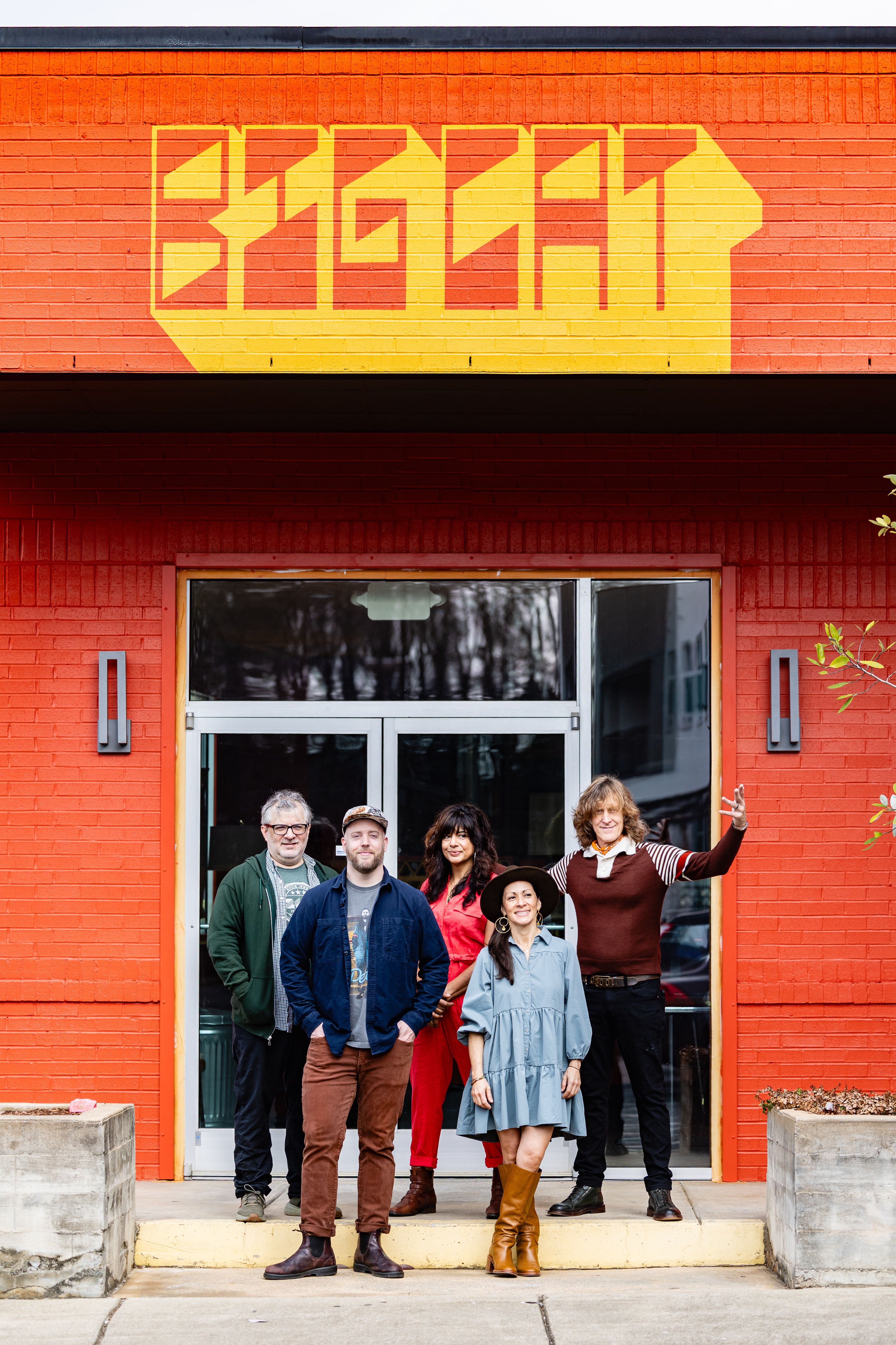 Four people stand in front of a red brick building with yellow geometric signage. They wear casual clothing including a blue dress with brown boots, red outfit, dark jacket, and brown sweater.