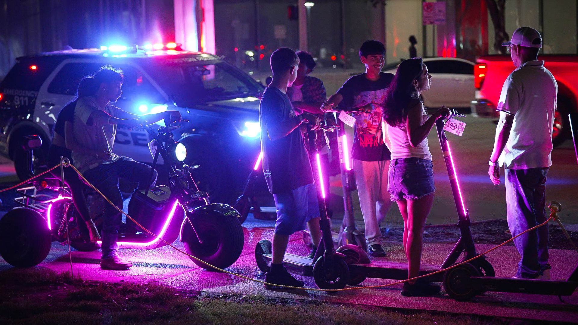 Young people mount e-scooters equipped with pink lights with a Houston police cruiser in the background