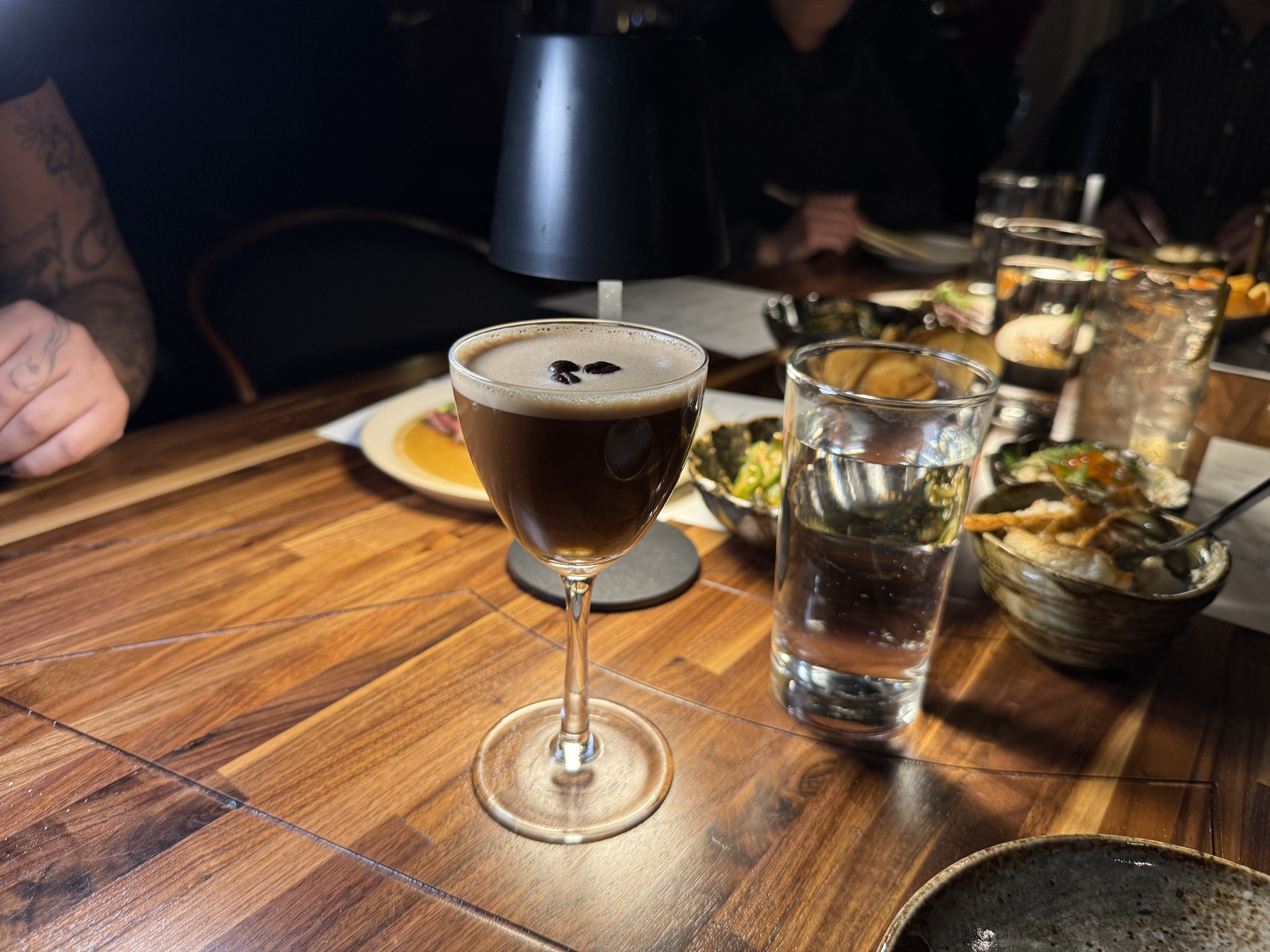 Espresso martini garnished with coffee beans in a glass on a wooden table next to a glass of water and various dishes with food under warm lighting.