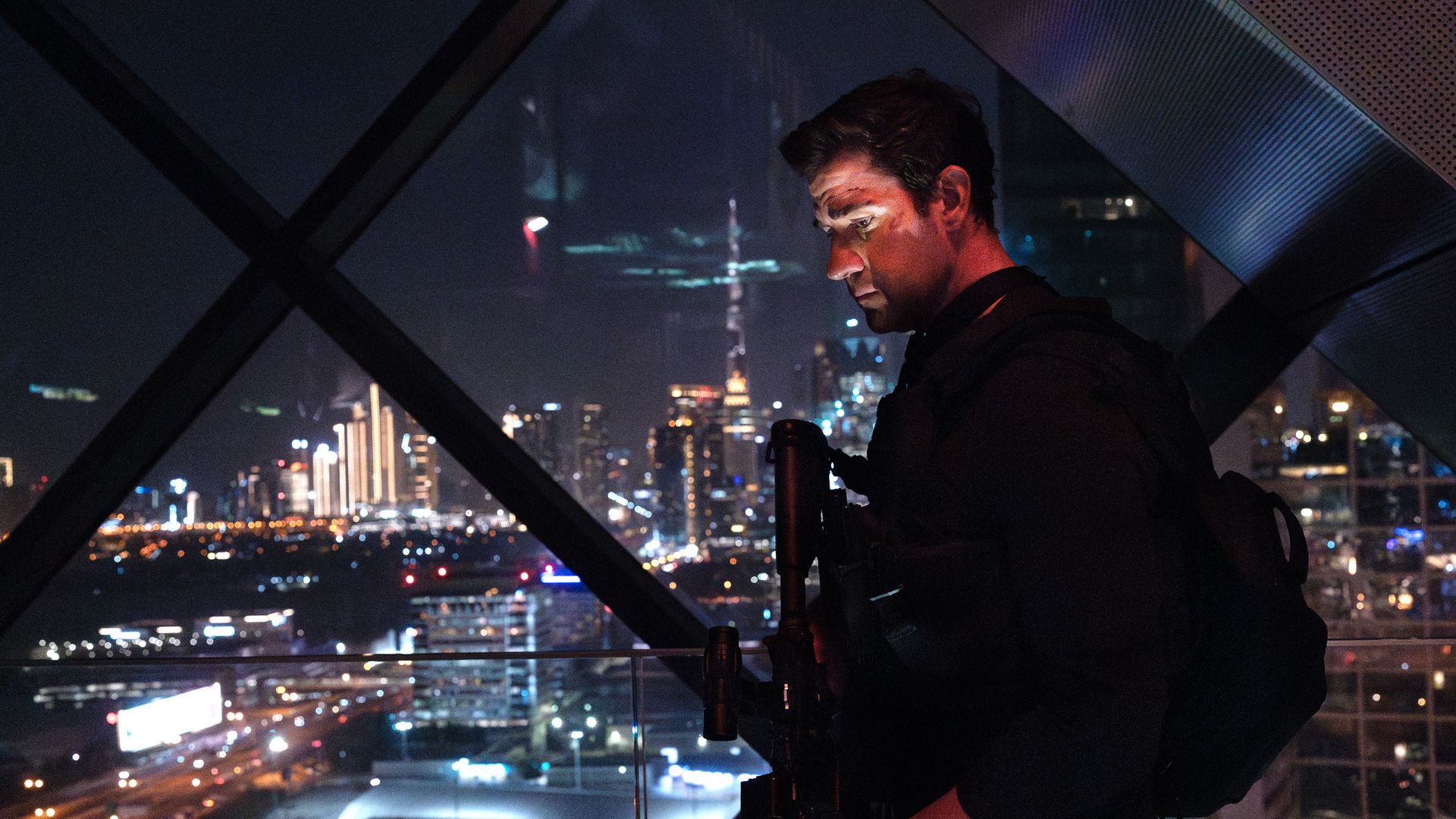 Night scene of a man in dark clothing, John Krasinski, leaning on a railing inside a glass-walled observation deck, gazing downward. City skyline with illuminated skyscrapers visible through diagonal metal frames.