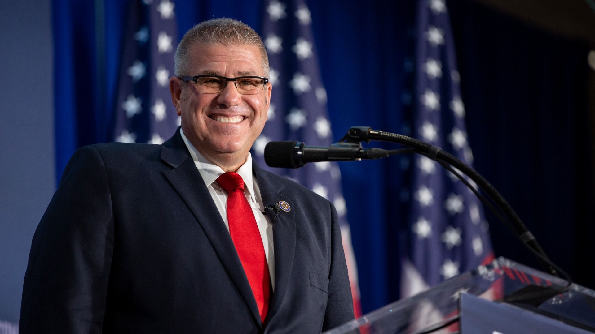 A smiling man in a dark suit and red tie speaks at a podium with a microphone, with American flags in the backdrop.