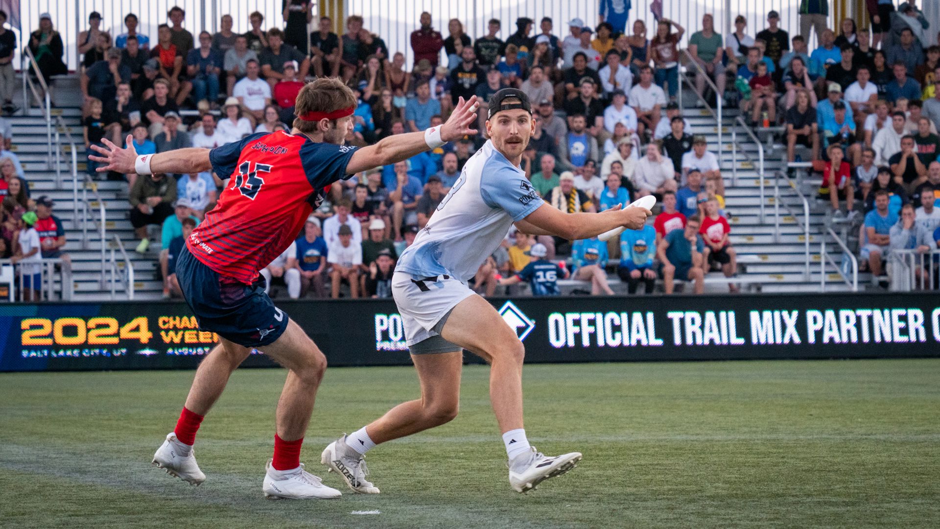 Two players on a green artificial turf field playing ultimate frisbee. One player has his arms spread-eagled attempting to block another one in a blue and white jersey, who's reeling back the disc preparing a throw.