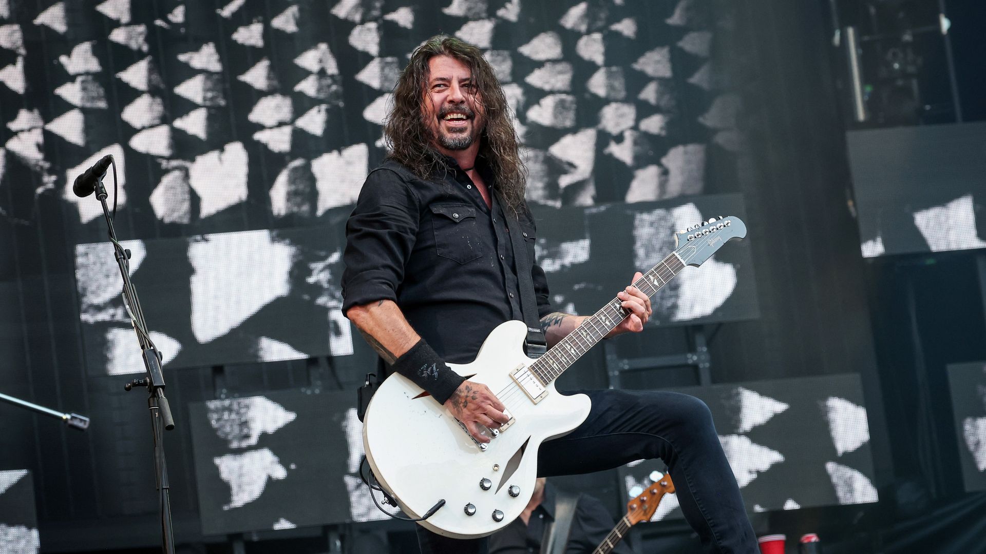 Male guitarist with long hair, wearing a black shirt, smiles while playing a white electric guitar on a concert stage with a black-and-white video backdrop and a microphone stand nearby.