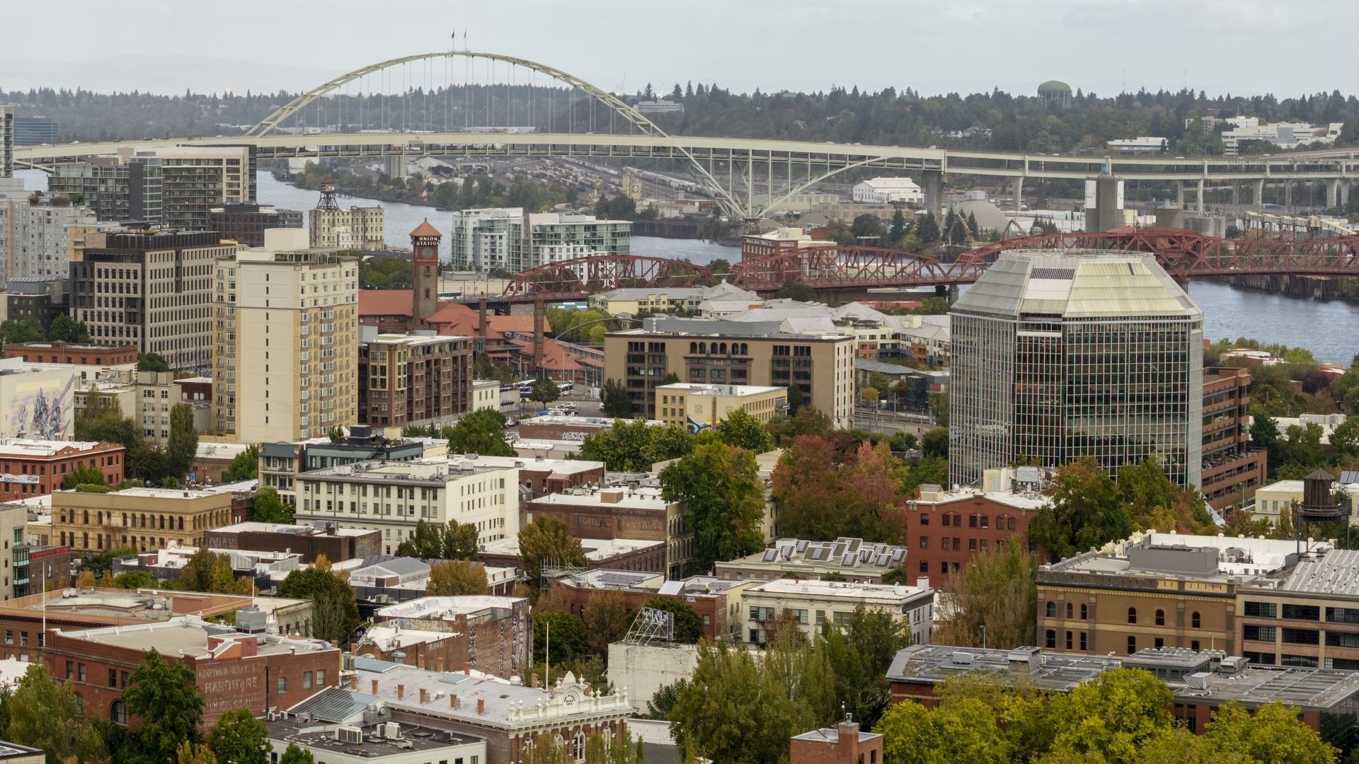 City riverfront scene with two bridges: a pale arched span in the distance and a red steel bridge nearby. Foreground shows trees, brick buildings, and a large glass office tower on the right.