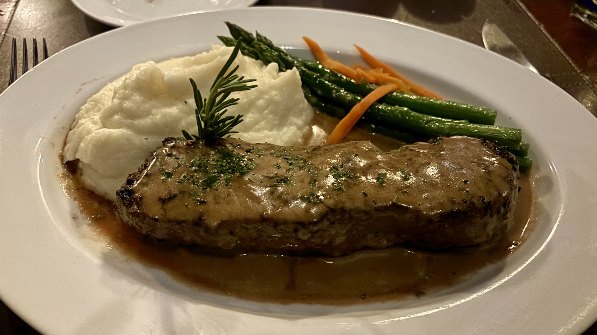 Plate with herb-crusted beef fillet in brown gravy, mashed potatoes topped with a rosemary sprig, and green asparagus with carrot ribbons.