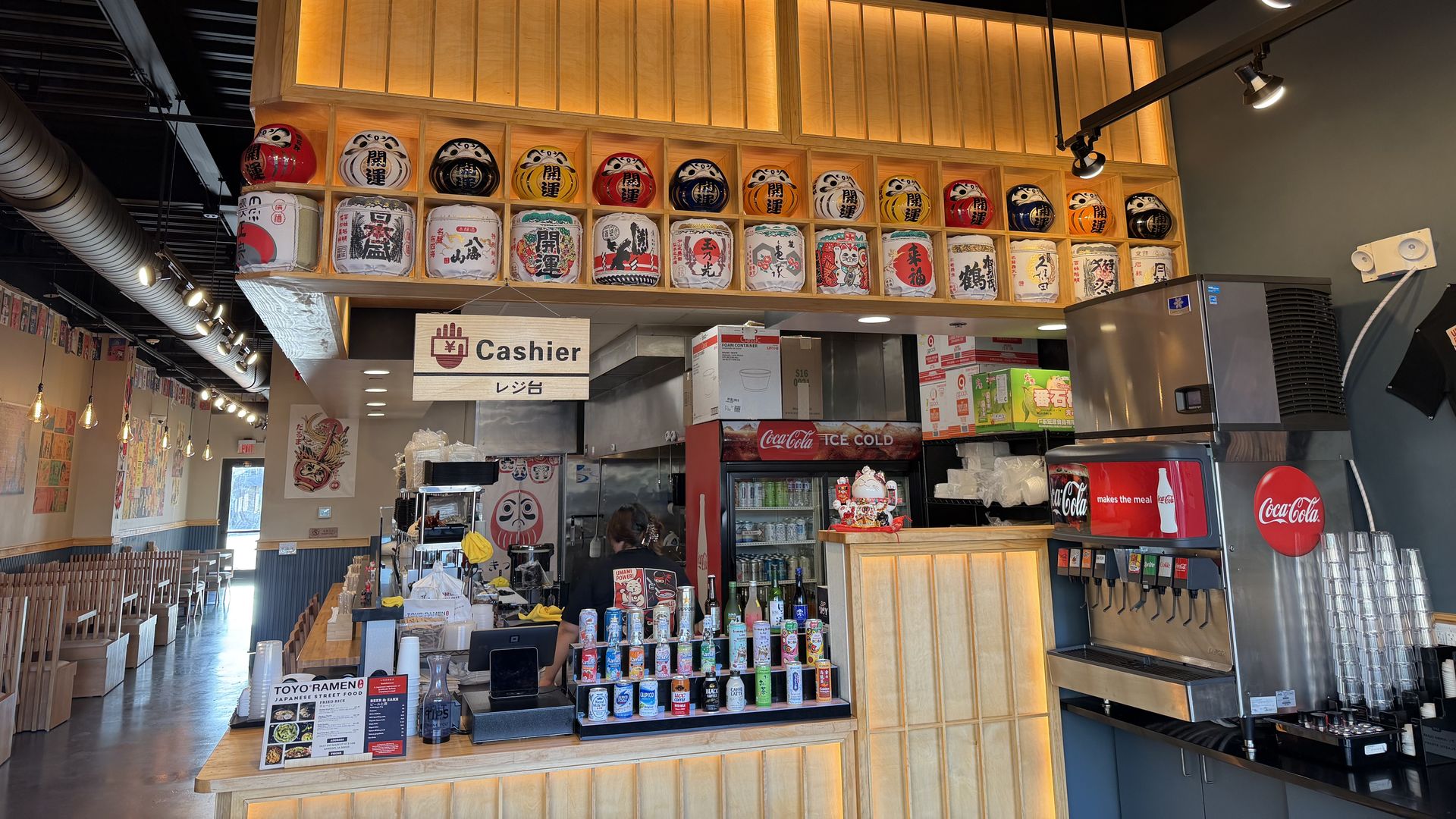 Interior of a Japanese ramen restaurant showing the cashier counter with sake barrels and daruma dolls displayed above, Coca-Cola drink dispenser, and wooden seating area with hanging lights