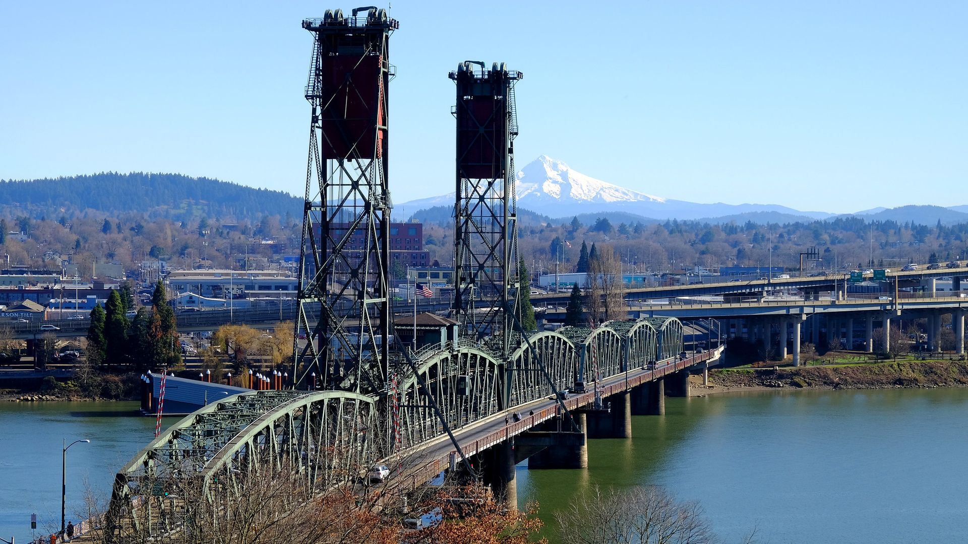 Steel truss bridge over calm river with twin vertical lift towers, cityscape, and snow-capped mountain in the background under a clear blue sky.