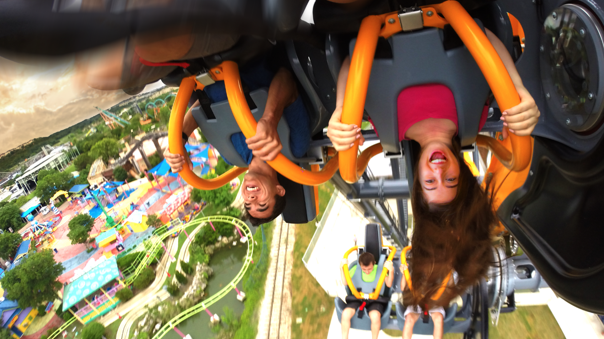 Two people upside down on an orange roller coaster at an amusement park, smiling and holding the bars tightly, with colorful rides and greenery below.