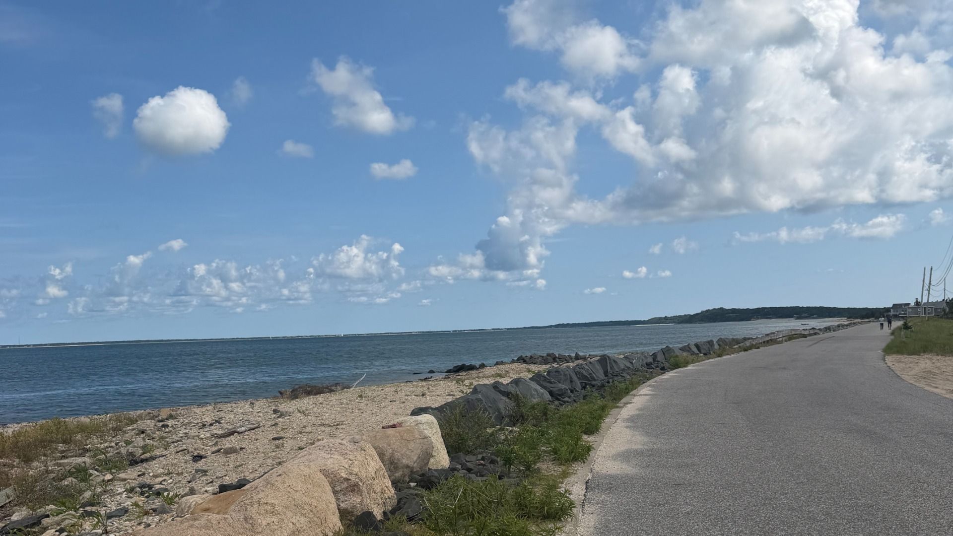 Coastal scene: a paved road runs along a rocky beach with blue water on the left and a cloudy sky above. Distant houses and power lines line the right, with a few walkers in view.