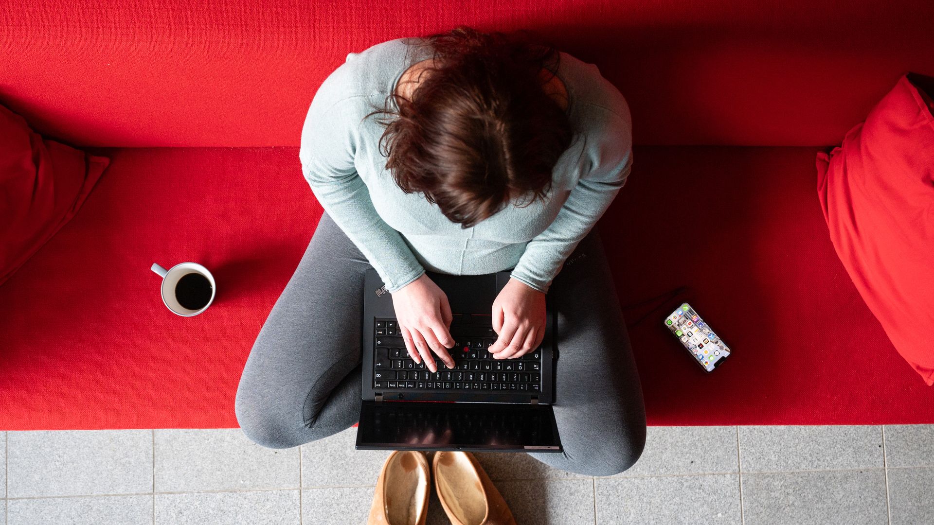 A person sits cross legged on a red couch with a laptop. 