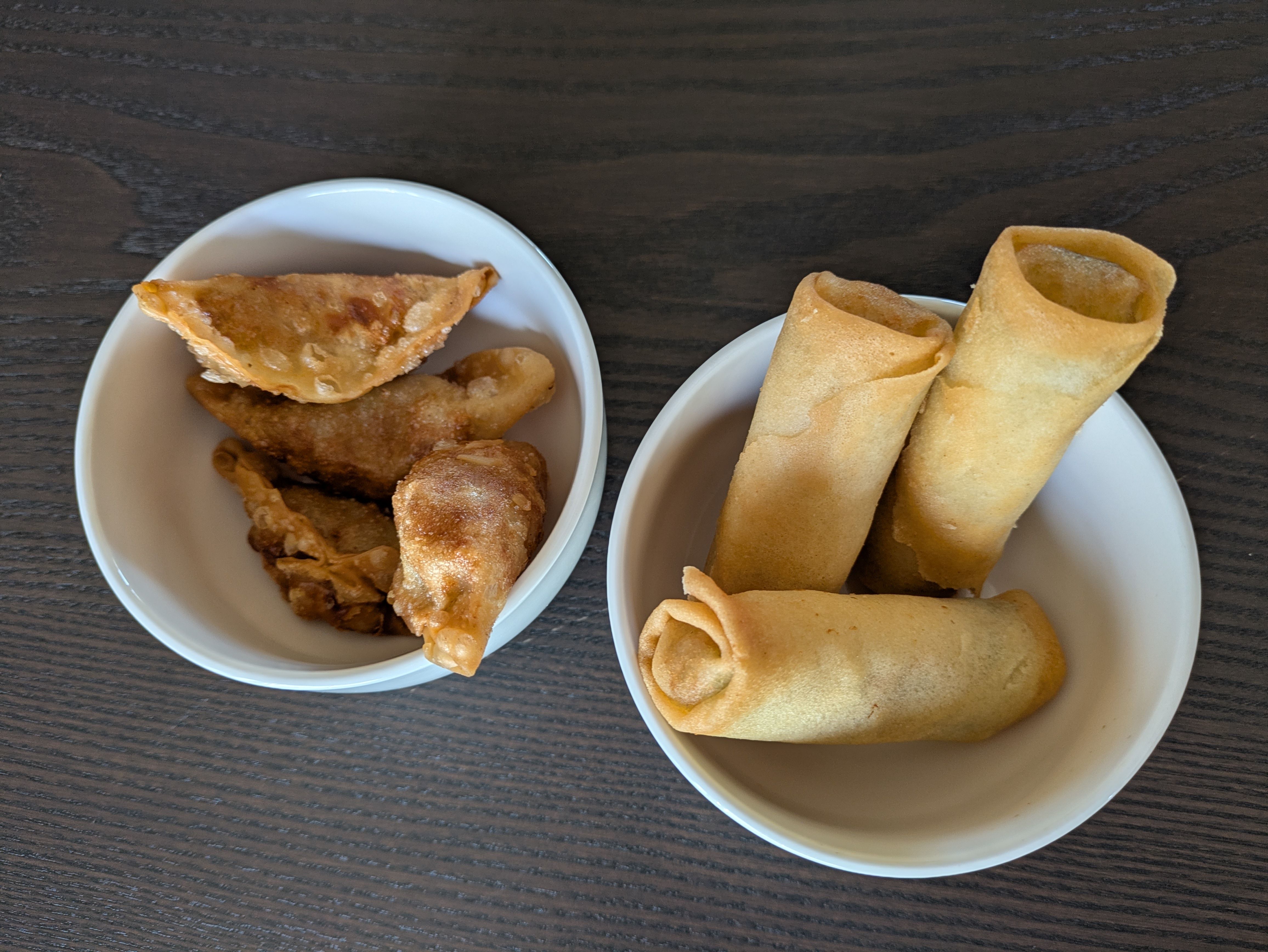 Two white bowls on a dark wooden table hold fried snacks: left bowl with golden-brown battered chicken pieces, right bowl with crispy rolled egg rolls.