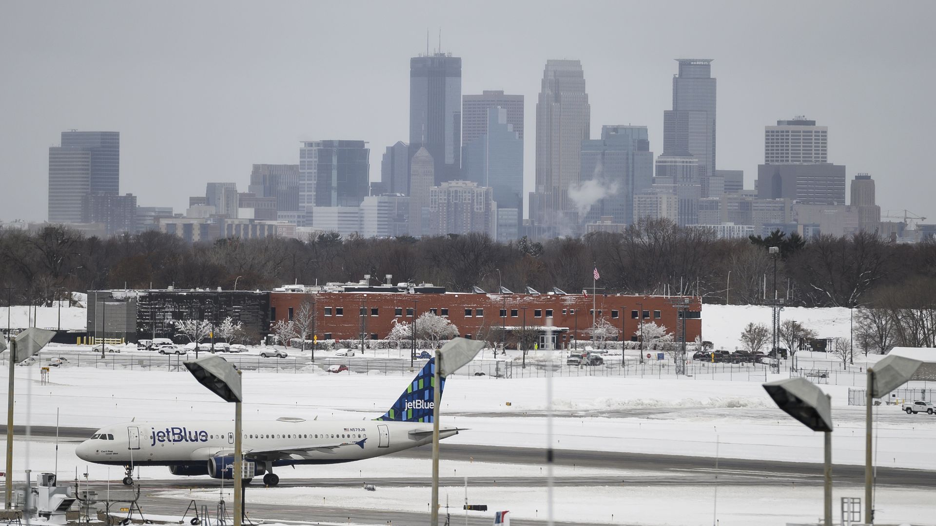 A white jetBlue airplane with a blue tail sits on the tarmac at an airport with a city skyline in the background.