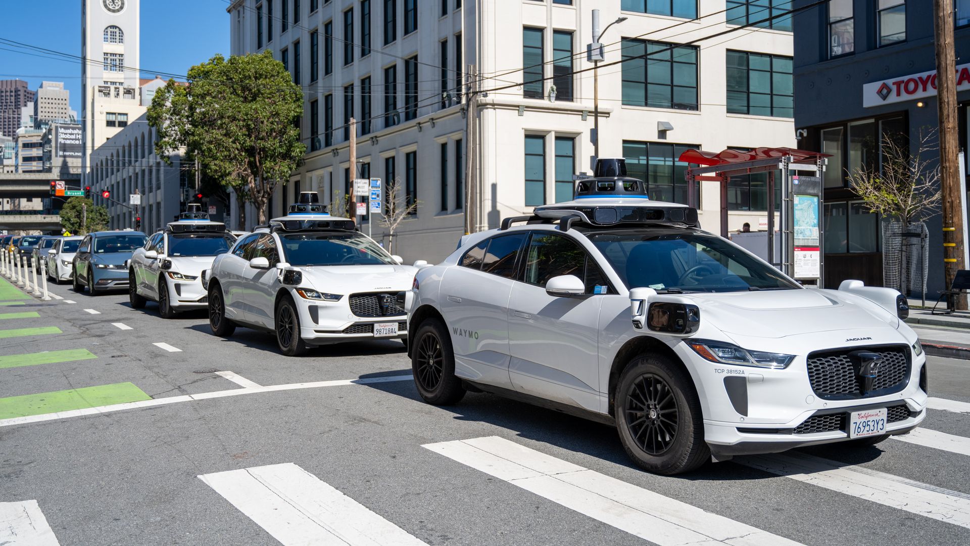 City street with a row of white Waymo autonomous taxis with roof sensors, lined up beside modern white office buildings; a tall clock tower on the left, blue sky, and power lines.