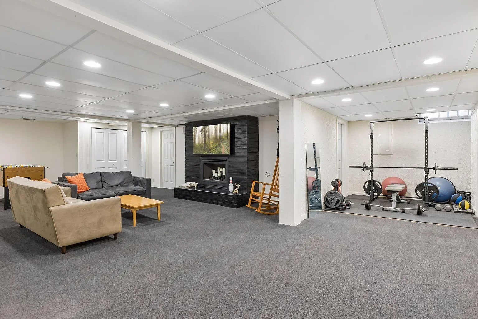 Spacious basement with white ceiling and recessed lights. Beige sofa and gray sectional around a coffee table near a black stone fireplace with a TV. Gym area has a squat rack, weights, and mirrors.