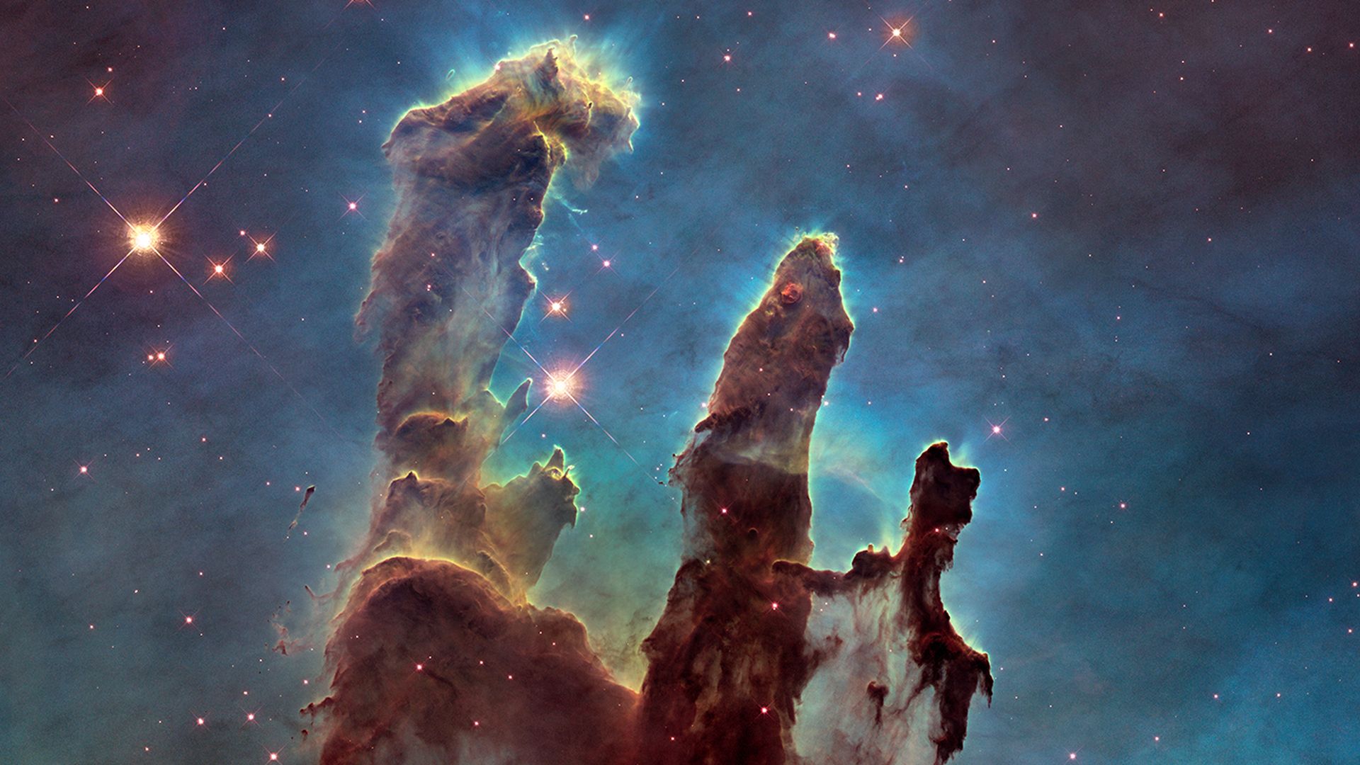 Pillars of gas and stars in brown on a background of blue and green light as seen by Hubble.