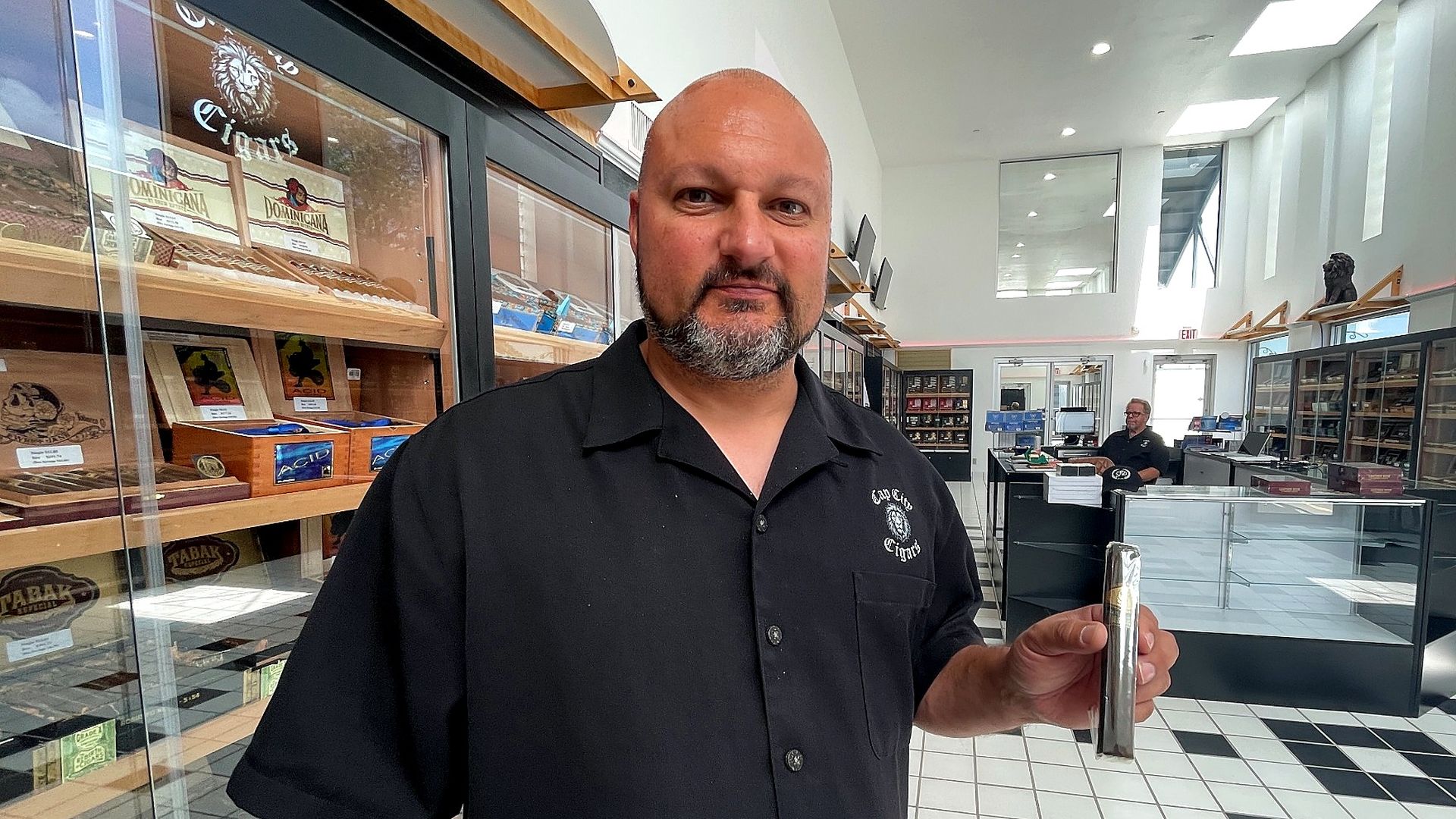 Smiling bald man with a gray beard in a black shirt stands in a cigar shop, holding a shiny metal object. Black-and-white floor, glass display cases, and shelves of cigars surround him.