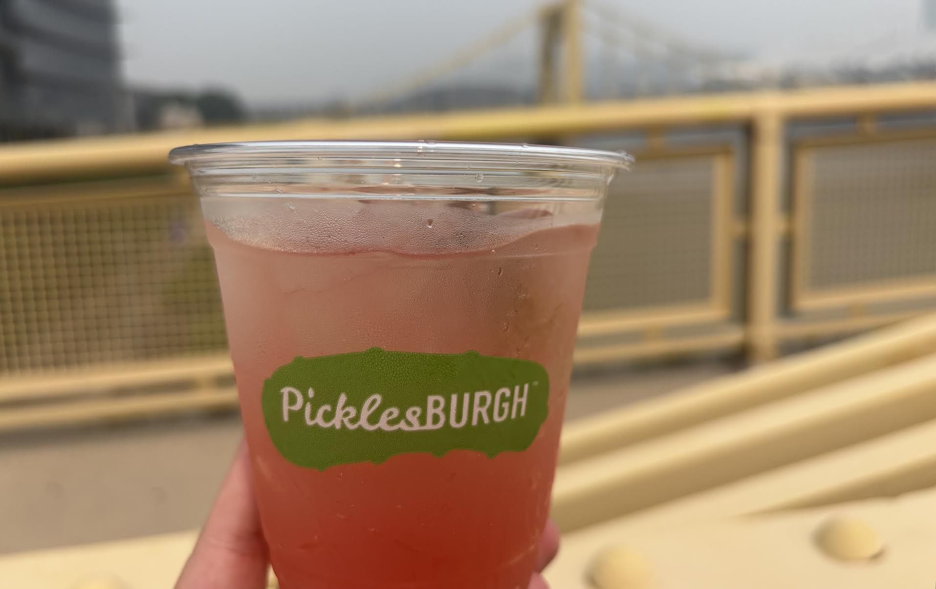 Close-up of a pink beverage in a clear plastic cup with condensation, held in front of a blurred railing and cityscape. The green label on the cup says "PicklesBURGH".