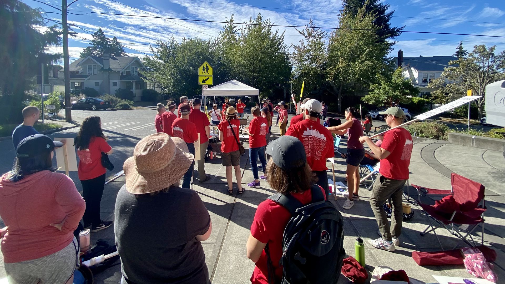 A group of people with signs and in red shirts stands on a sidewalk listening to someone speak. 