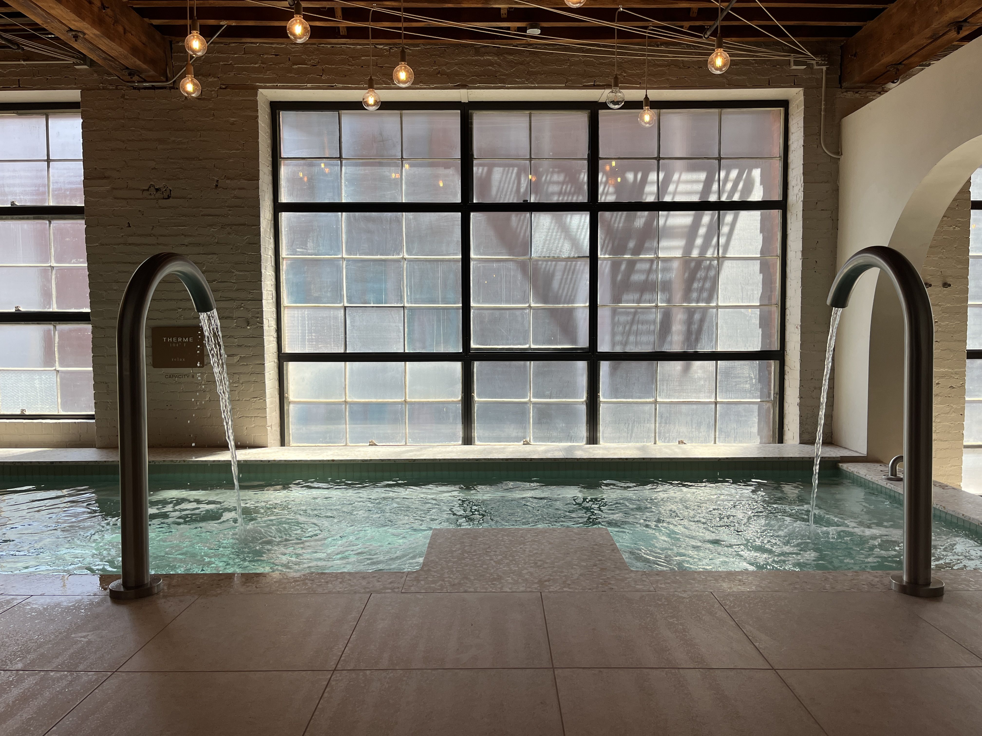 Indoor pool with two curved water fountains pouring water, large window with frosted glass panels, exposed brick walls, wooden ceiling beams, and hanging light bulbs.