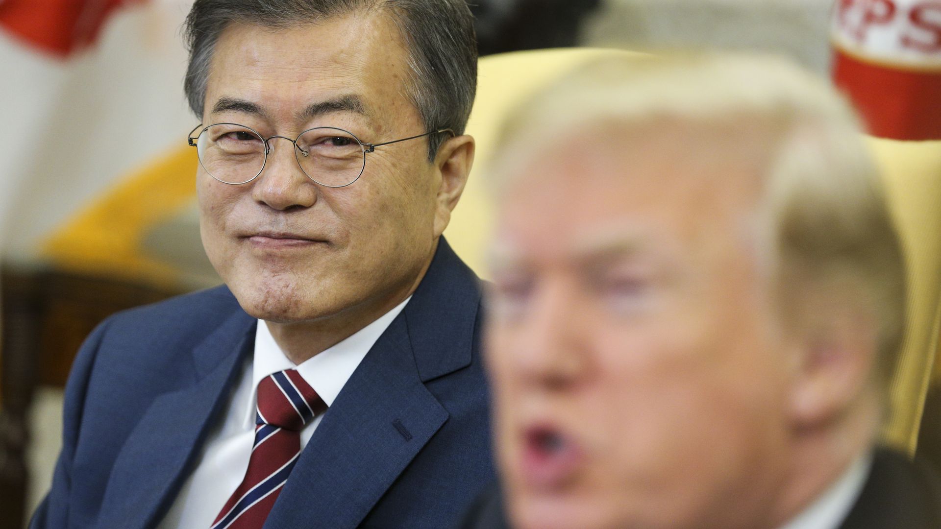 Moon Jae-in sits looking sideways at Trump, who is blurry in the foreground while speaking.