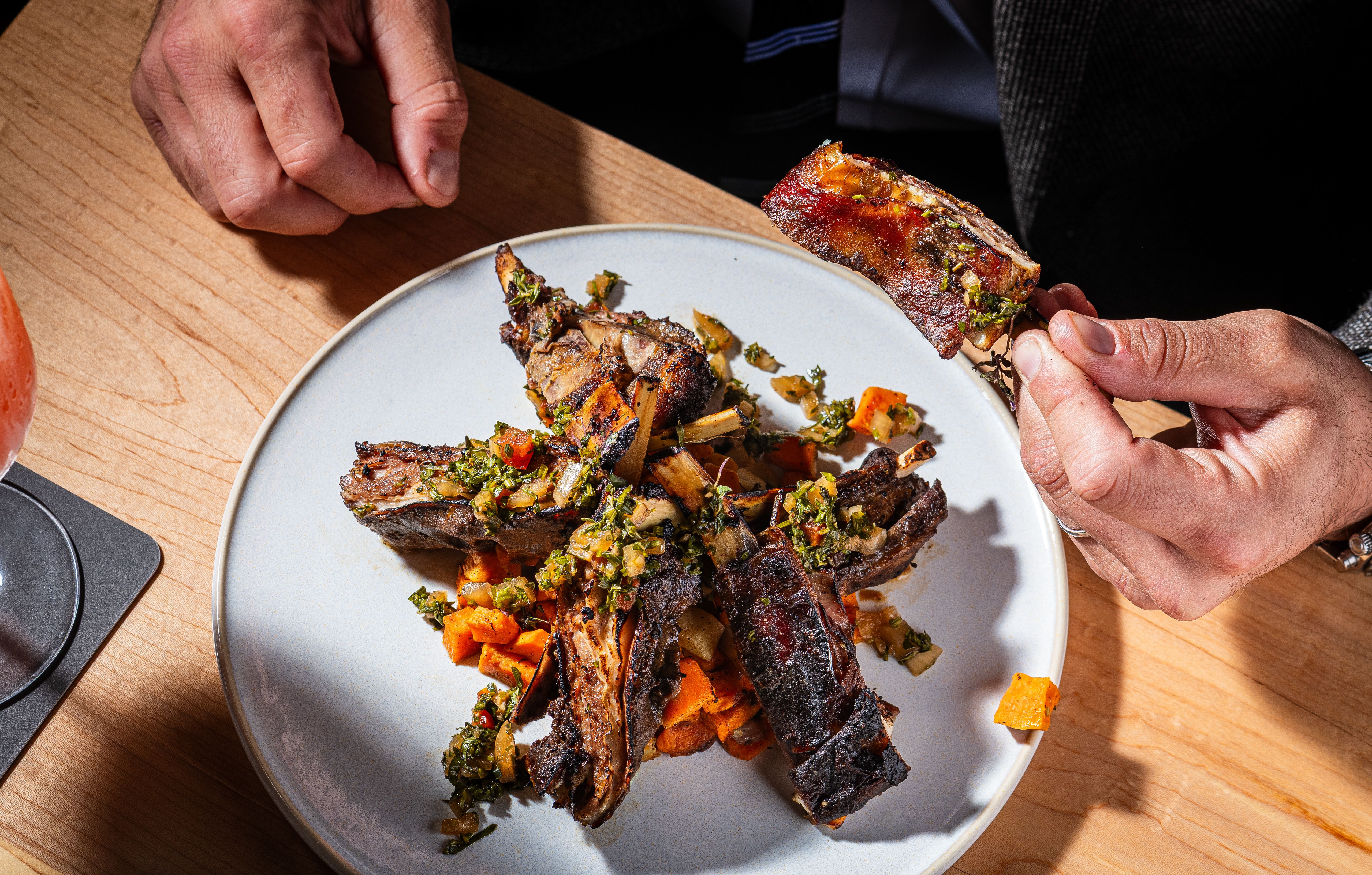 Close-up of two hands on a wooden table; one holds a rib over a light-gray plate of grilled ribs topped with chopped herbs, onions, and diced orange vegetables.