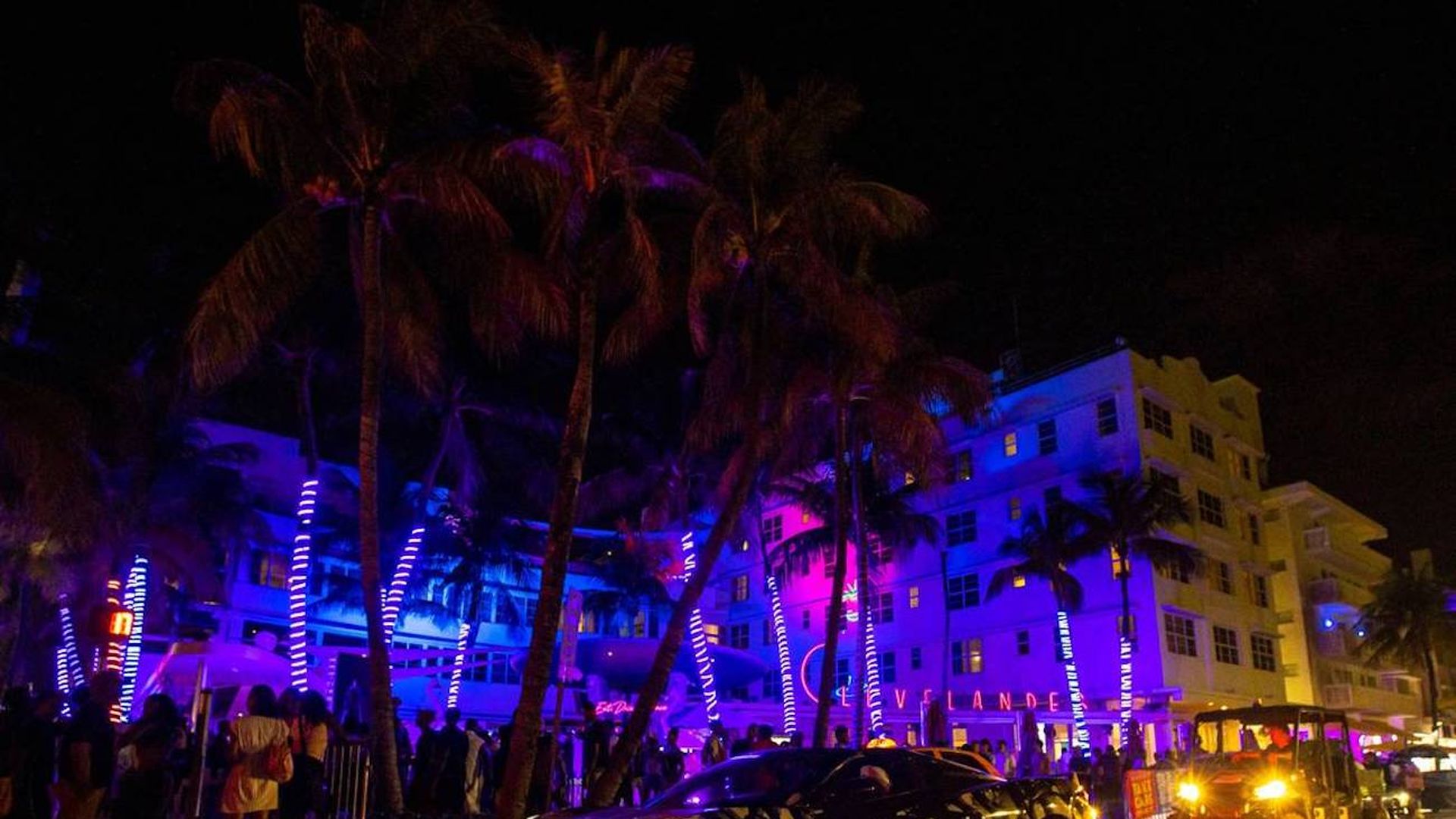A building is lit up in blue light beside palm trees and a crowd and a sportscar.