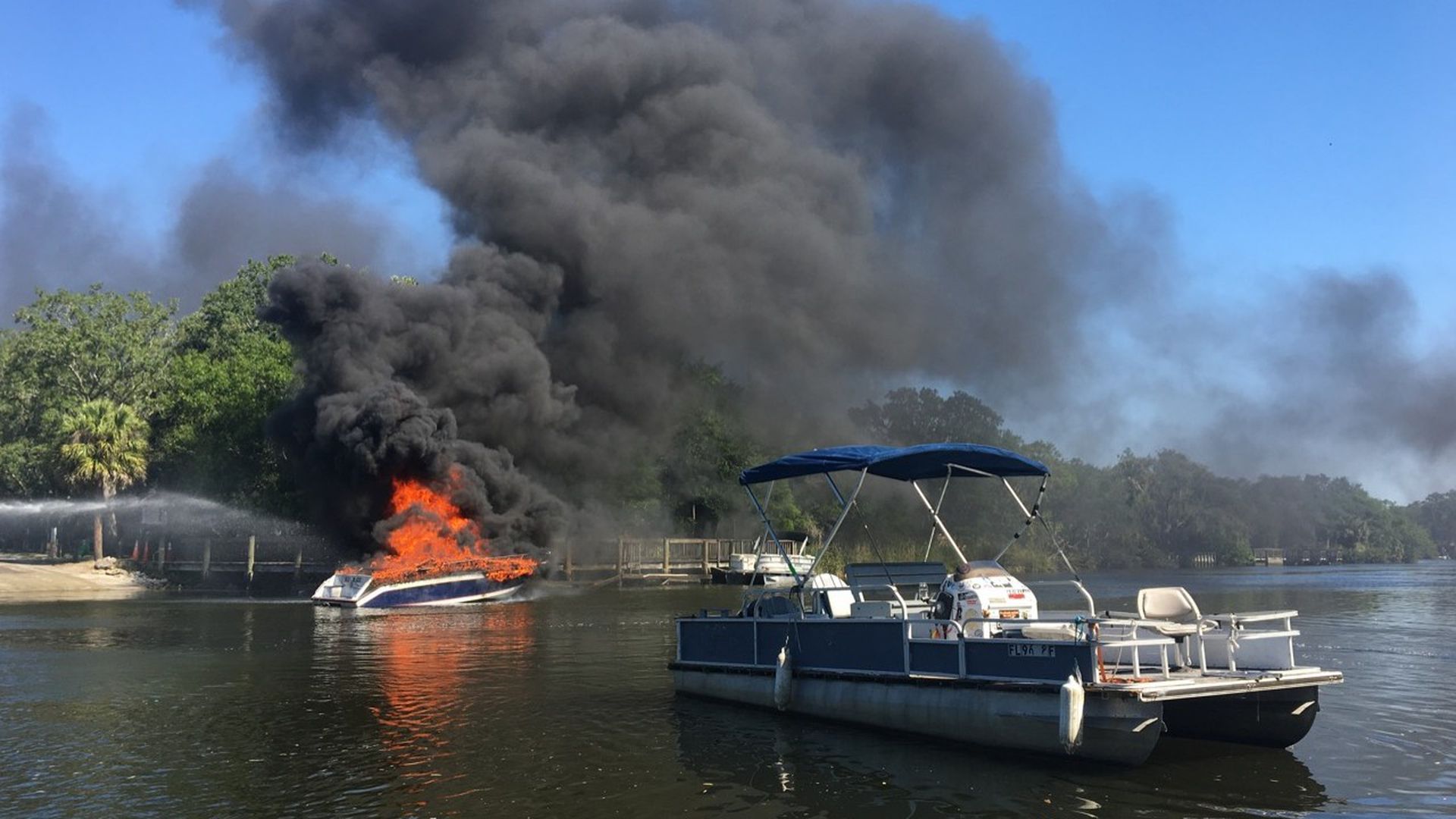 A boat up in flames on the water, with black smoke billowing overhead and another boat nearby