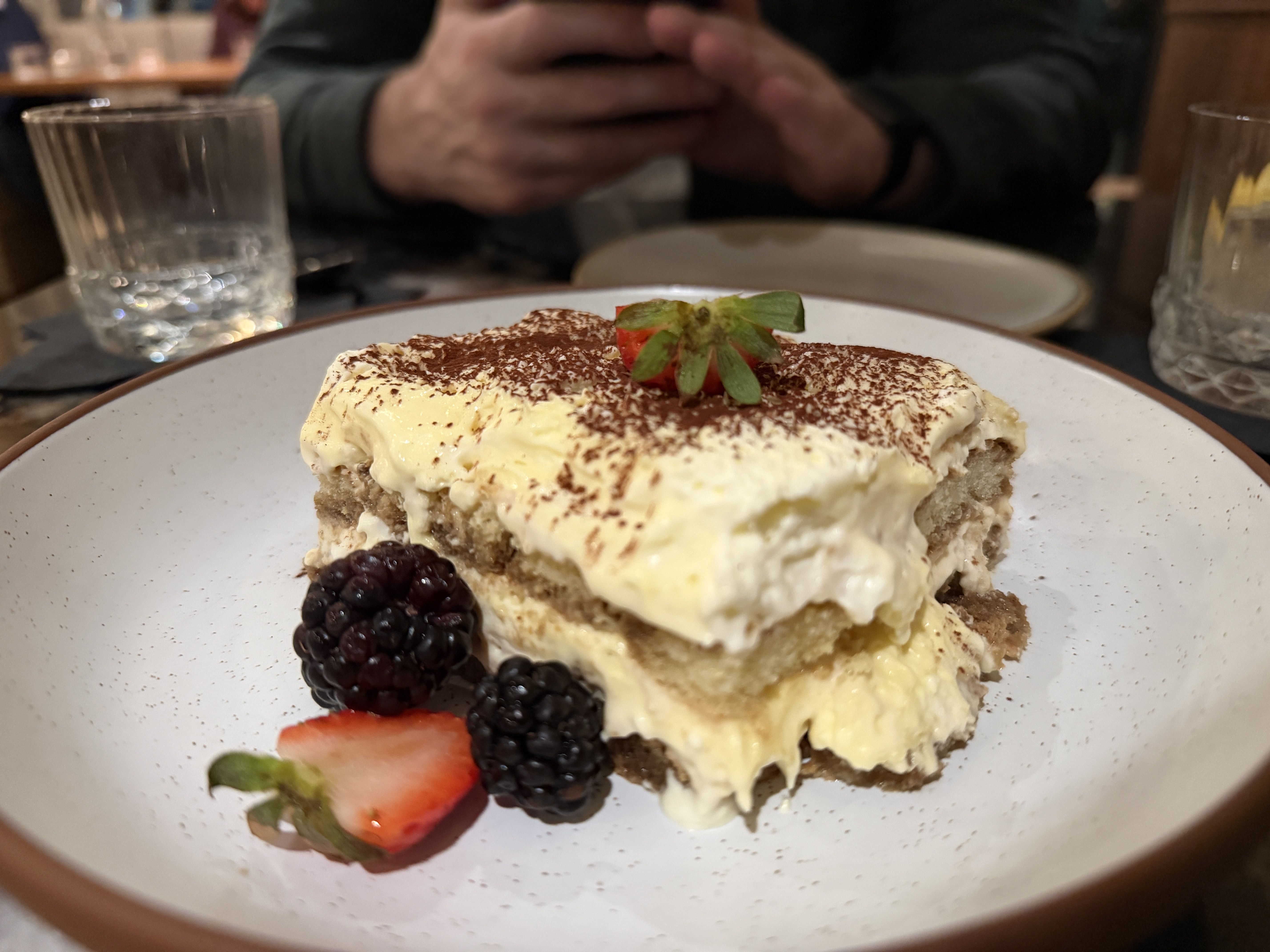 Slice of creamy tiramisu dessert topped with cocoa powder and a strawberry half, served with blackberries and a strawberry slice on a white plate with brown rim.