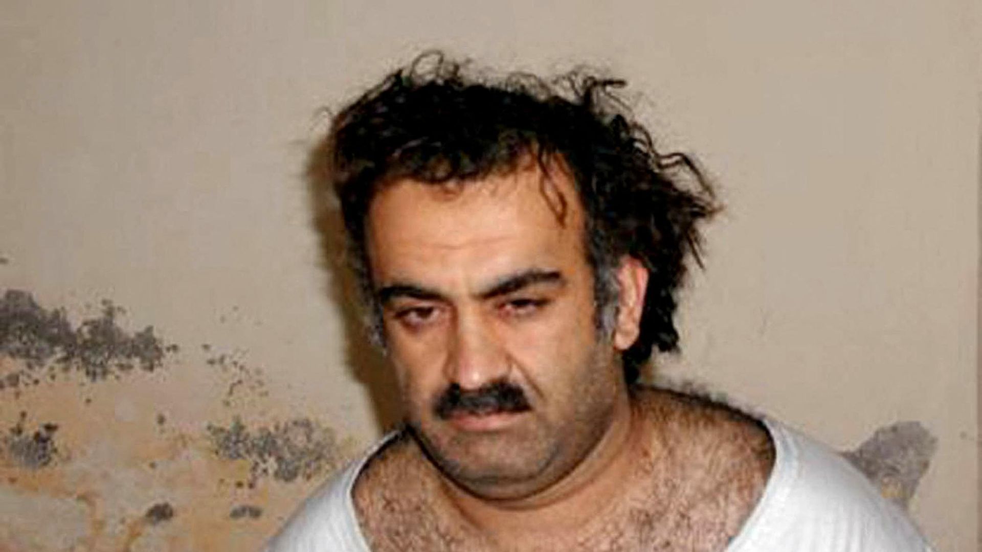 Man with dark, messy hair and a mustache wearing a white tank top, looking at the camera with a neutral expression, in front of a beige, stained wall.