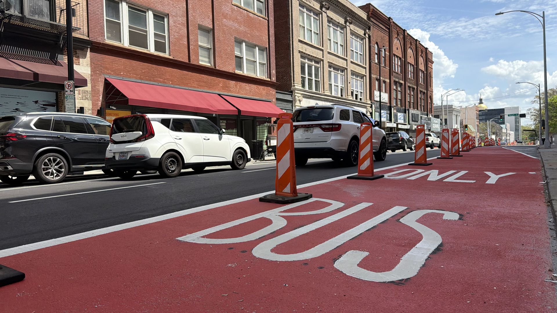 City street with a red bus-only lane marked "BUS," orange construction barriers, parked cars along the curb, brick storefronts with red awnings, and a blue sky with clouds.