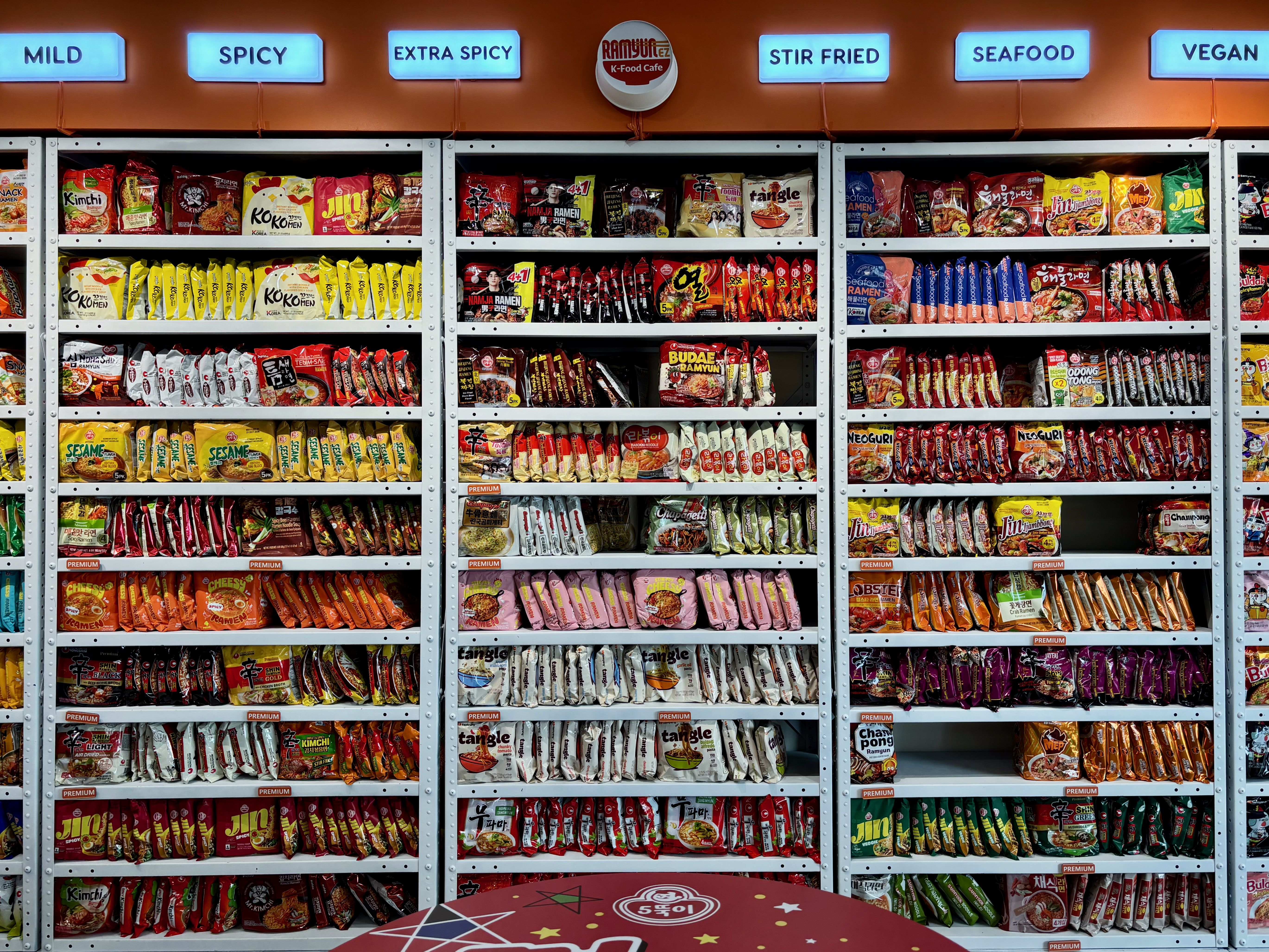Wide display of instant ramen bundles on gray metal shelves, organized by heat level: MILD, SPICY, EXTRA SPICY, STIR FRIED, SEAFOOD, VEGAN. Colorful packets densely lined across shelves.