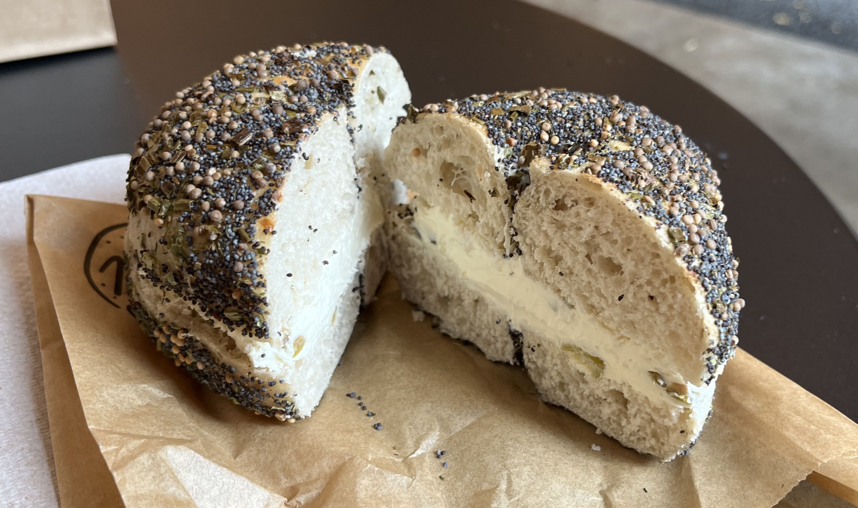Two halves of a Chicago bagel with visible poppy seeds, mustard seeds, and herbs, spread with cream cheese, placed on brown wax paper on a dark surface.
