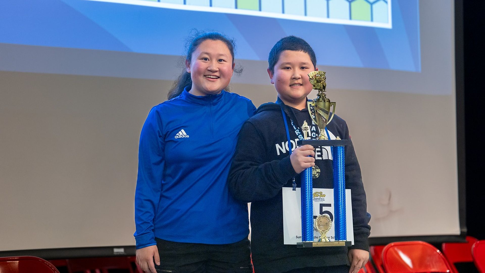 Two smiling kids stand on a stage; a girl in a blue Adidas jacket stands beside a boy holding a tall blue trophy with a gold cup, while a hexagonal-patterned screen looms behind.