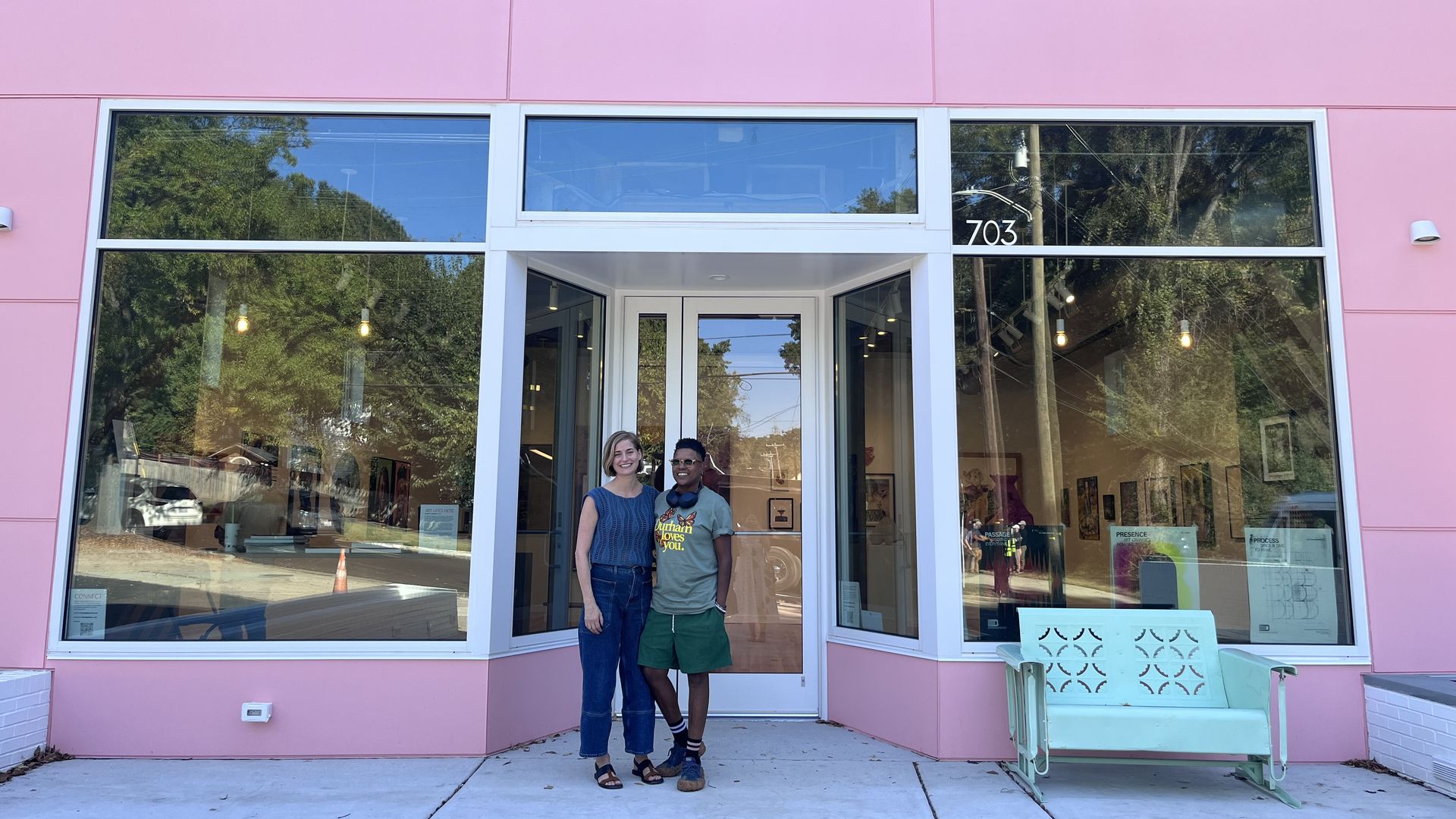Two people stand smiling in front of a pink storefront with large glass windows and white frames. A glass door entrance shows the number 703; a white outdoor bench sits to the right.