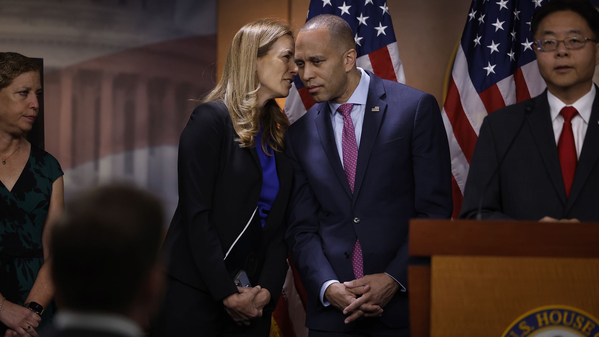 Reps. Mikie Sherrill whispers to Rep. Hakeem Jeffries while both wear suits and stand with their arms clasped in front of a row of American flags and next to colleagues speaking at a press conference.