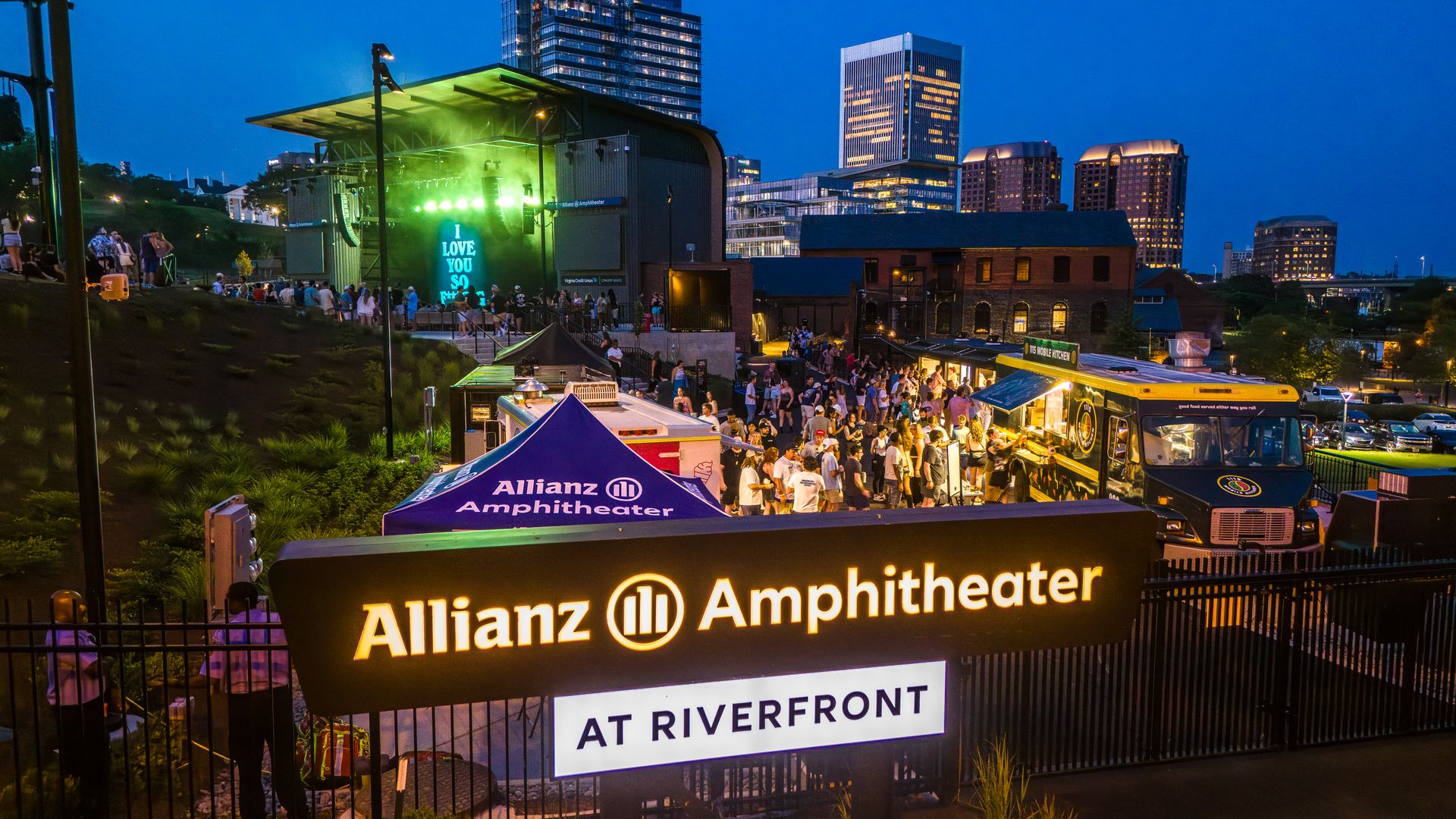 Blue-hour view of Allianz Amphitheater: a green-lit outdoor stage, cheering crowd on the lawn, tents and stalls, with a skyline of tall buildings in the evening sky.