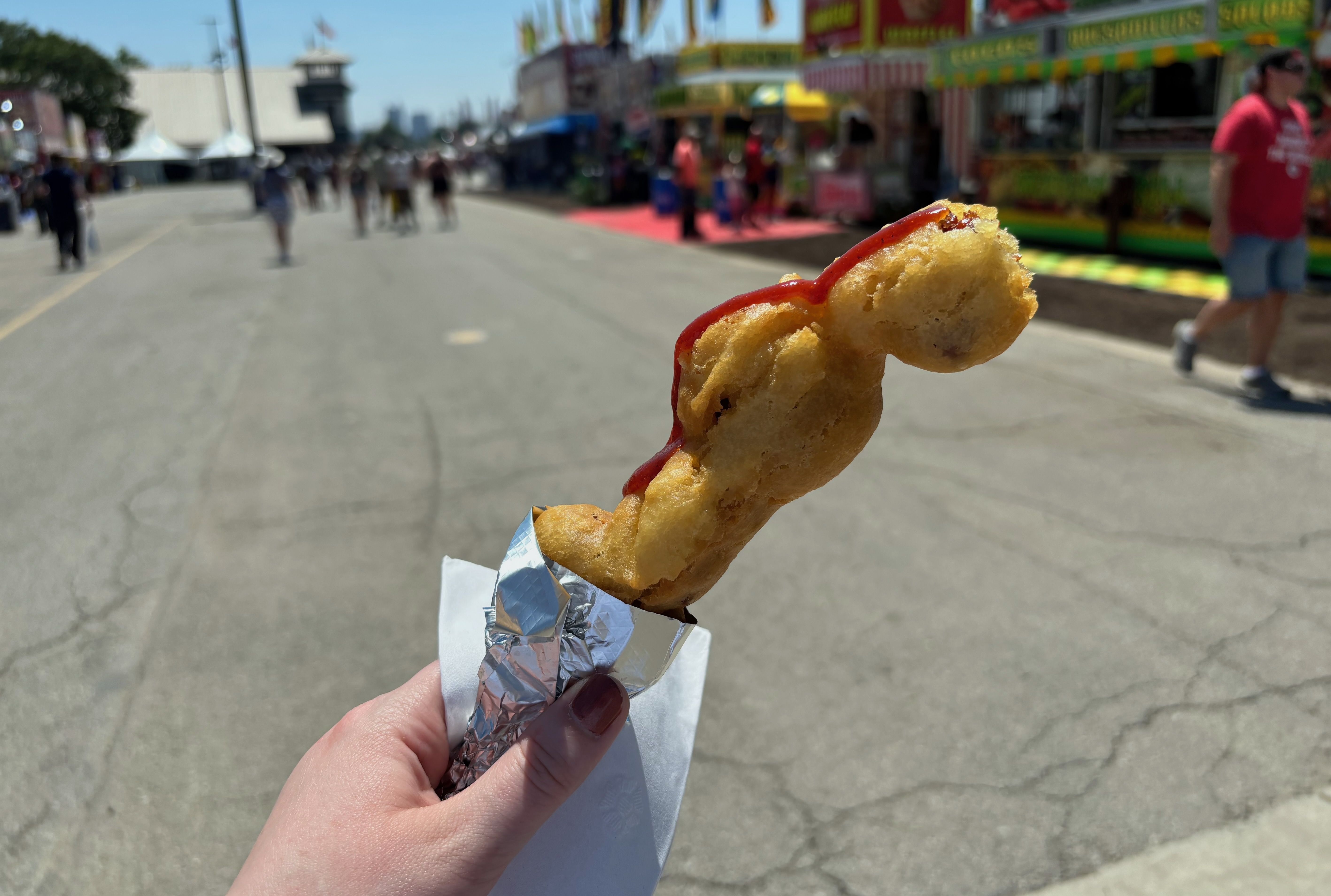 Hand holding a deep-fried food item with barbecue sauce on top, wrapped in foil and a napkin, at an outdoor fairground with food stalls and people in the background.