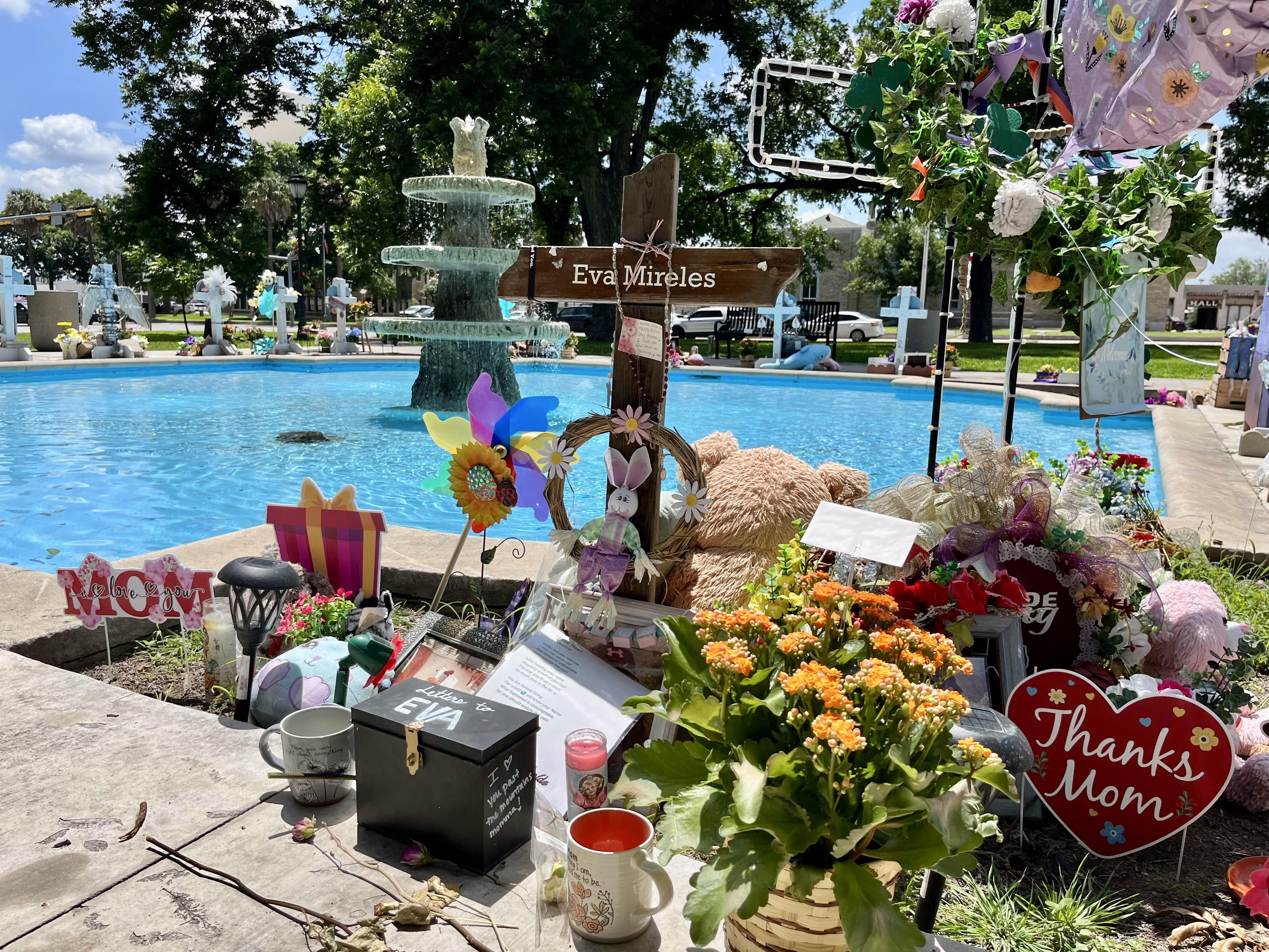A cross for Eva Mireles stands again a bright blue fountain in a square in downtown Uvalde, where other crosses sit around the pool to honor victims of the mass shooting at Robb Elementary School.