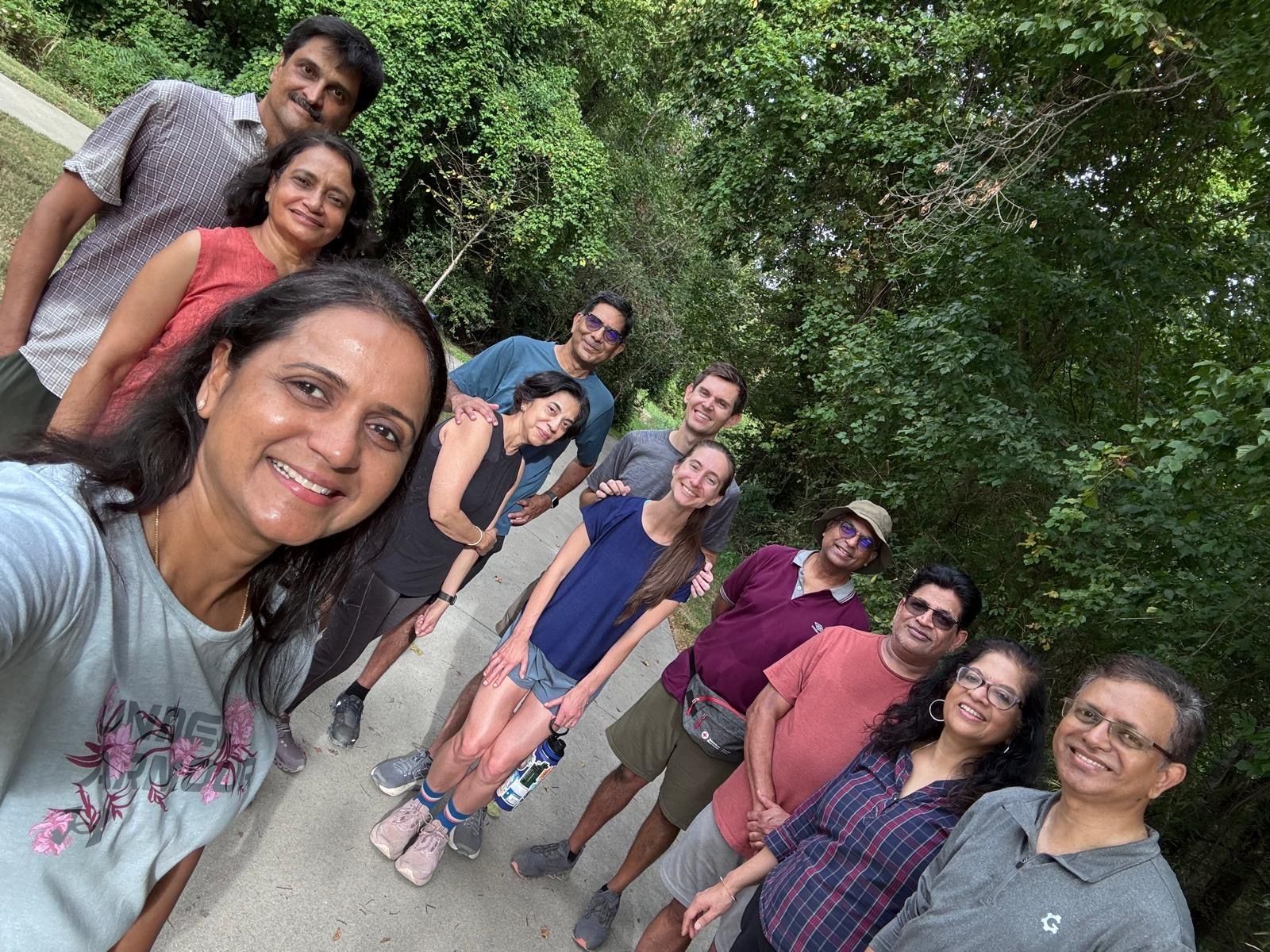 Selfie of a diverse group of friends on a sunny forest trail. Woman in a light gray shirt smiles in the foreground; others stand behind her in casual outfits, with lush green trees all around.