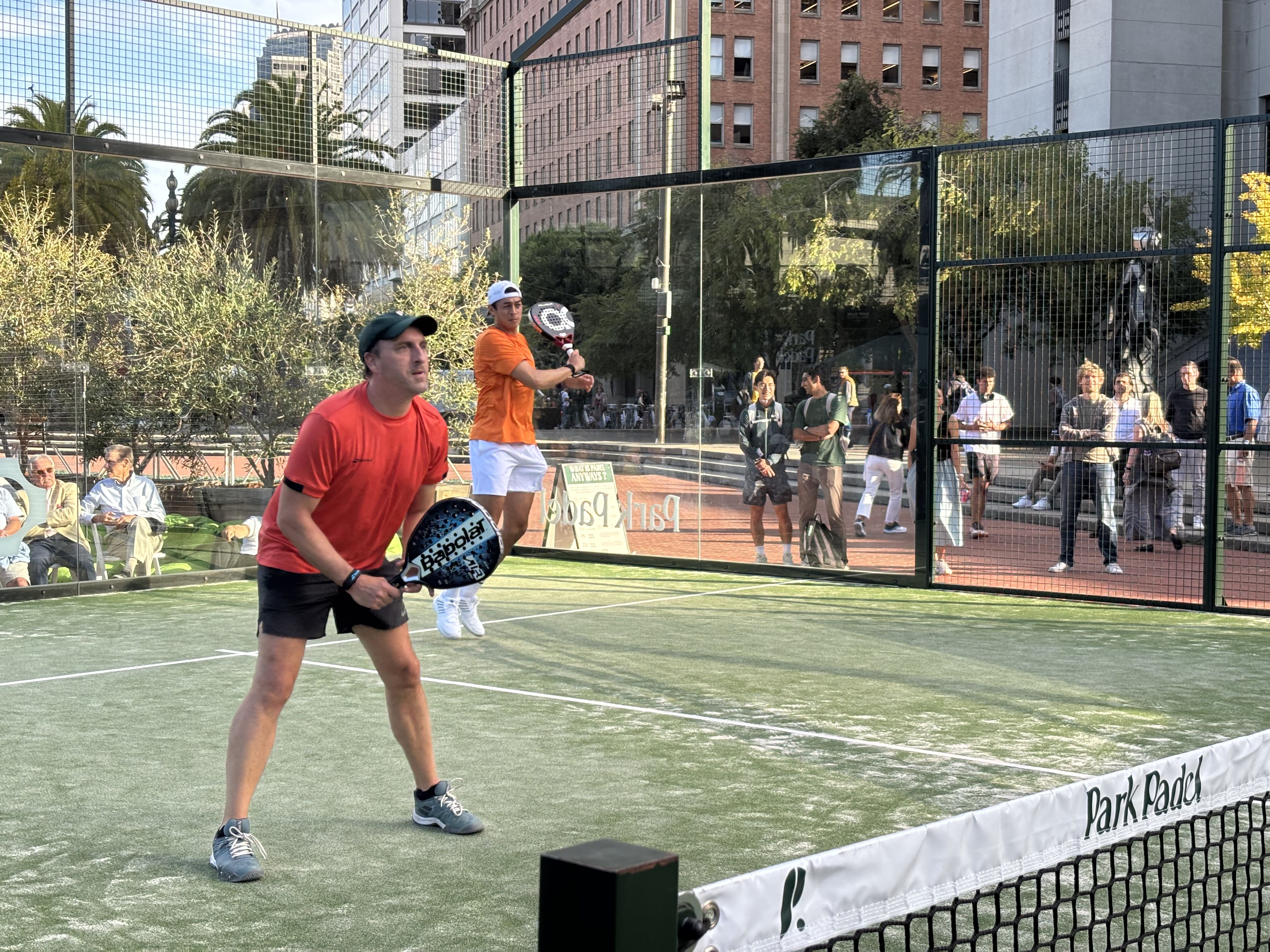 Two men playing padel on green court surrounded by glass walls in urban park; one in red shirt and black shorts, the other jumper in orange shirt and white shorts, crowd watching beyond.