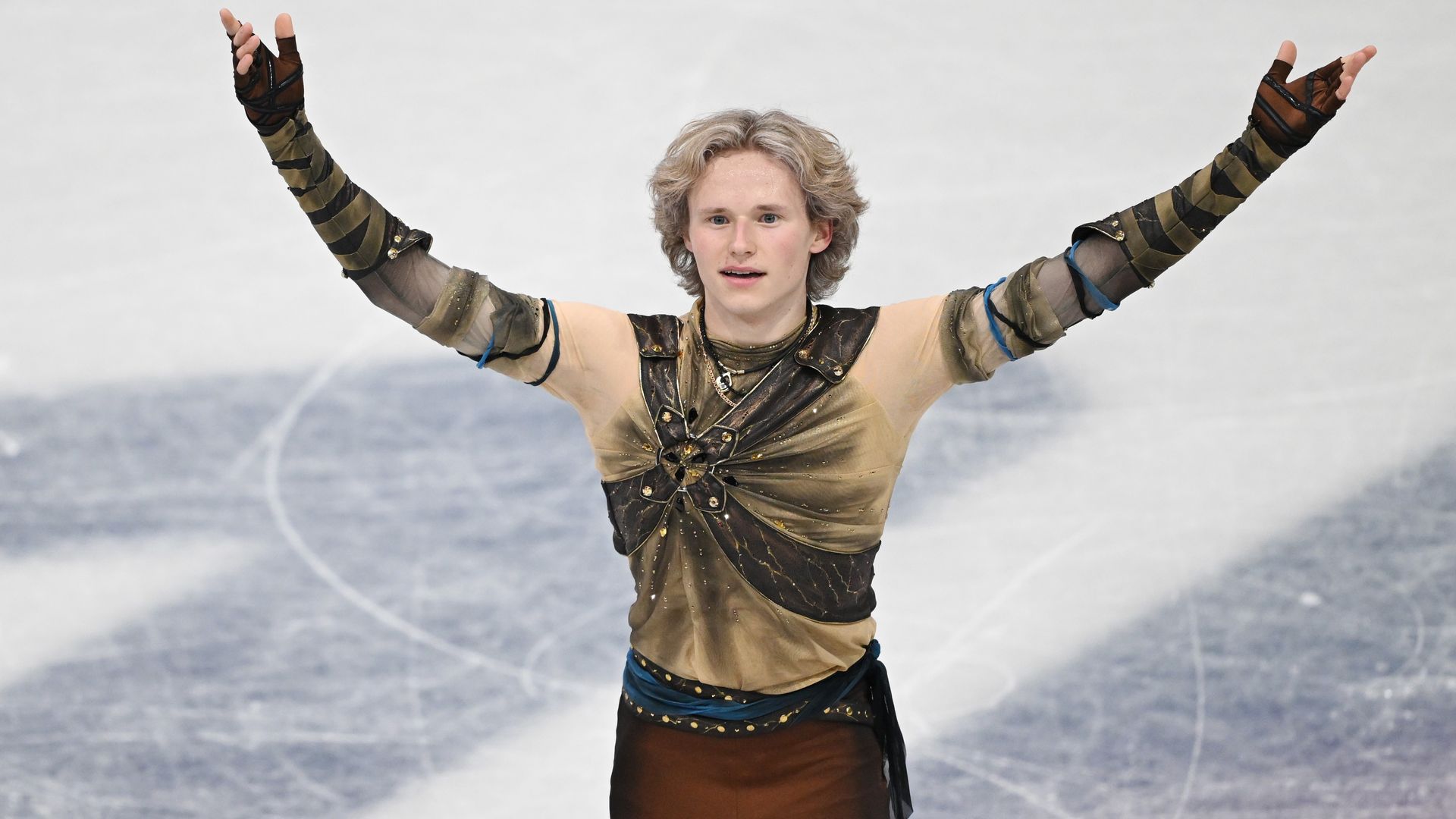 Figure skater Ilia Malinin in brown and beige costume with arms raised, standing on ice rink with visible skate marks.