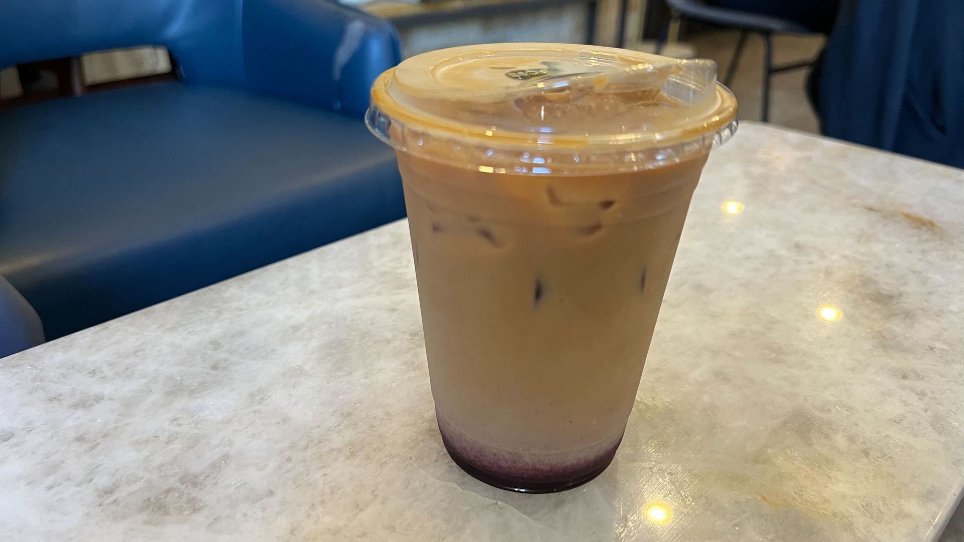 Blueberry iced coffee in a clear plastic cup with a lid on a white marble table, featuring a dark purple layer at the bottom