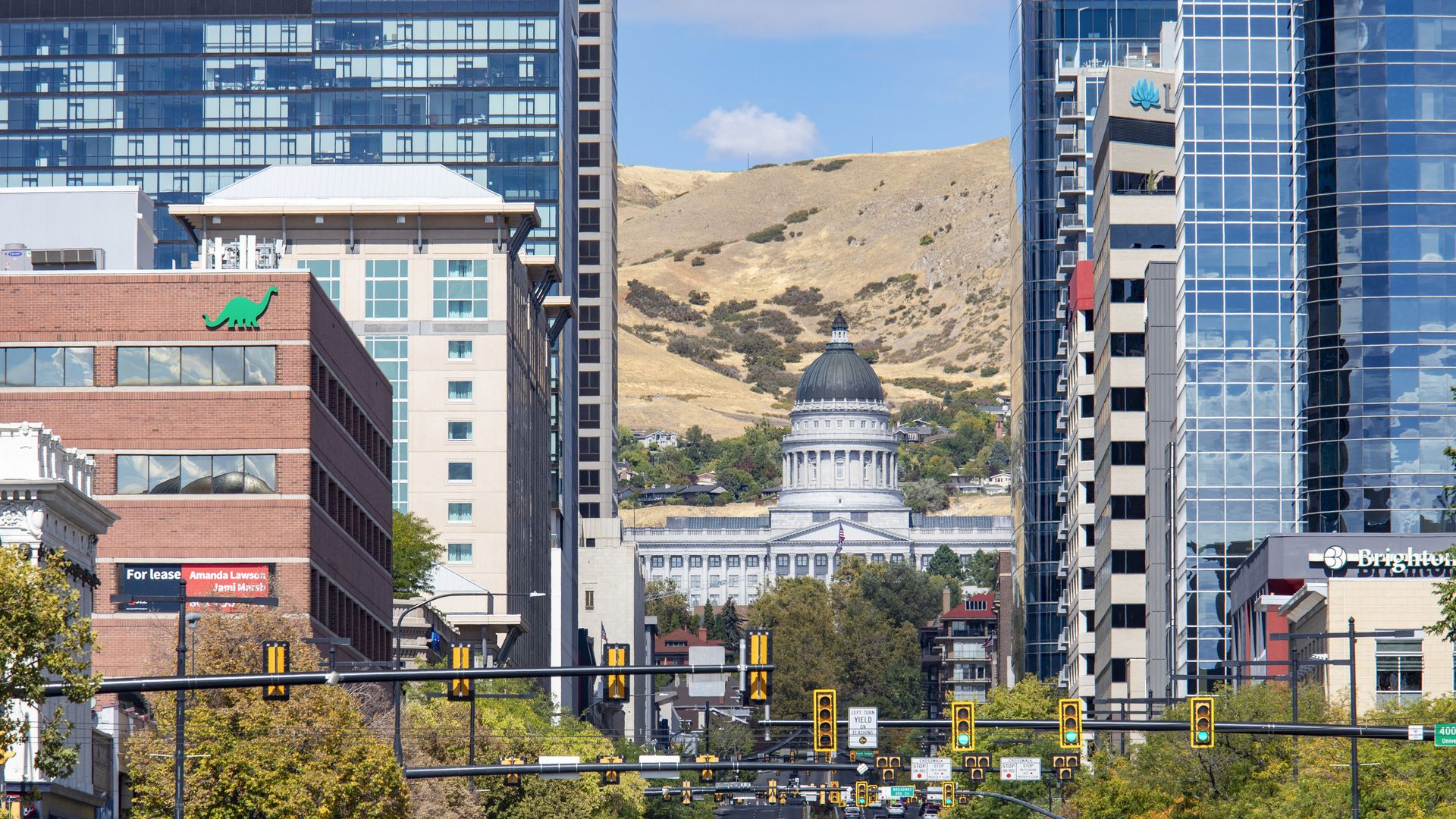 Downtown city street with traffic, framed by tall modern buildings and greenery, leading to a neoclassical capitol building with a dome, hills in the background under a blue sky.